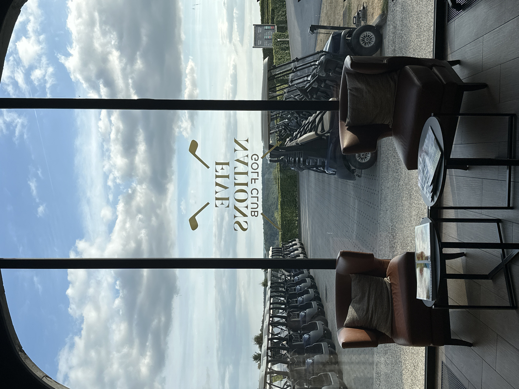 The image shows a view through a window, likely from a golf clubhouse in Méan, Belgium. The scene is set on a bright, cloudy day, possibly late morning or early afternoon.
The main subjects are golf carts parked in neat rows outside, viewed from an interior lounge area. Inside, two comfortable brown leather armchairs with cushions are visible, placed near small dark tables. One table has a magazine or brochure on it. The interior flooring appears to be made of wood planks.
Through the window, beyond the golf carts, there's a landscape with greenery and a sign visible in the distance. The sky is filled with dramatic, large white clouds against a light blue background.
Reflected on the window, there is text that reads "FIVE NATIONS GOLF CLUB" in gold lettering, with a golden golf club symbol to the left. The text is reversed due to the reflection. The overall impression is one of a modern, comfortable space offering a view of the golf course.
