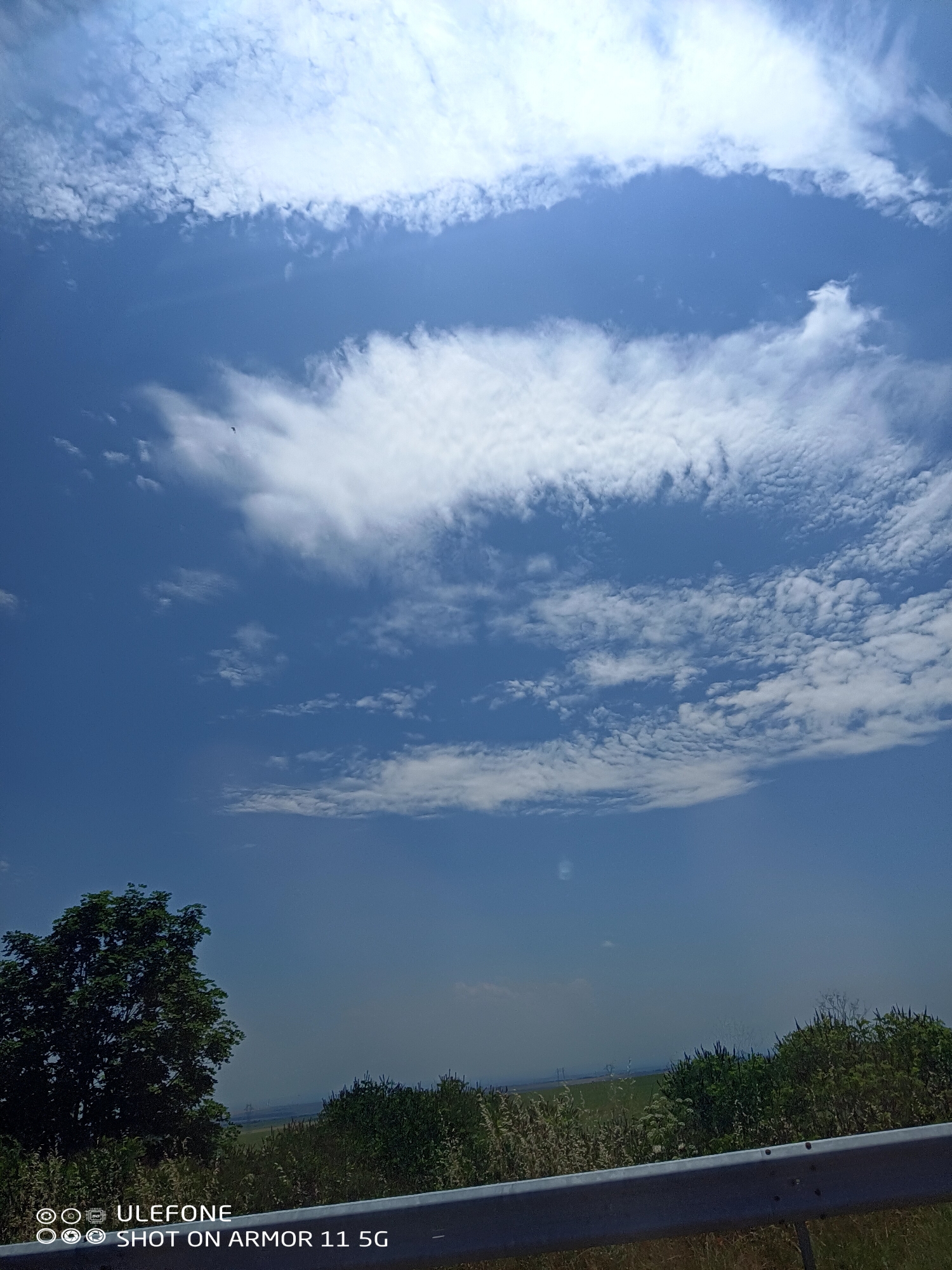 The image displays an outdoor daytime scene, likely taken from a vehicle, in the city of Unknown, Unknown. The upper two-thirds of the frame are dominated by a clear blue sky interspersed with various formations of white clouds. A prominent band of layered stratocumulus or altocumulus clouds extends horizontally across the middle of the sky. Above this, a larger, more diffuse cloud mass is present in the upper left, while the upper right features distinct, brighter cumulus clouds. A small, dark, winged object, potentially a bird or drone, is discernible in the upper-middle left sky, above the horizon. The lower third of the image shows a rural or semi-rural landscape. On the far left, a large, dark green, leafy tree stands prominently. To the right of the tree, the distant horizon reveals hazy green fields and indistinct, light-colored structures, possibly industrial or power infrastructure. In the foreground, dense green bushes and lighter, dry vegetation occupy the right side. A silver metal guardrail with visible fastening points extends horizontally across the very bottom of the frame, partially obscuring the immediate foreground vegetation. The lighting suggests a bright, sunny day. No people or overt actions are depicted. The bottom left corner contains a digital watermark stating "ULEFONE" and "SHOT ON ARMOR 11 5G".