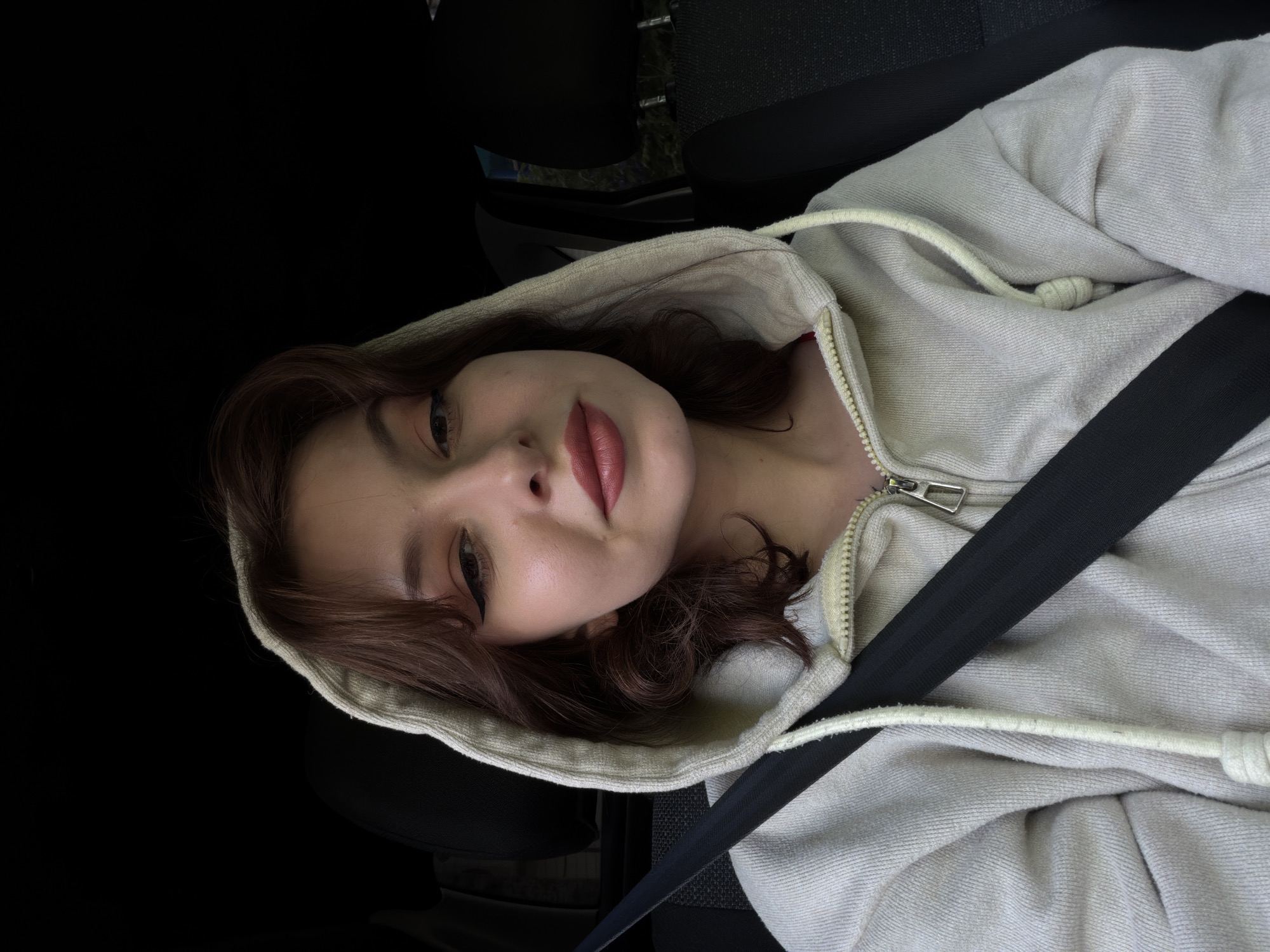 A young woman with shoulder-length brown hair, wearing a light-colored zippered hoodie, is seated in a car, likely in the driver's seat, indicated by the visible seatbelt across her chest. She is looking directly at the camera, with her head slightly tilted. Her makeup includes winged eyeliner and lipstick. The background is dark, suggesting the interior of a vehicle, with a hint of the outside world visible through a window on the right.