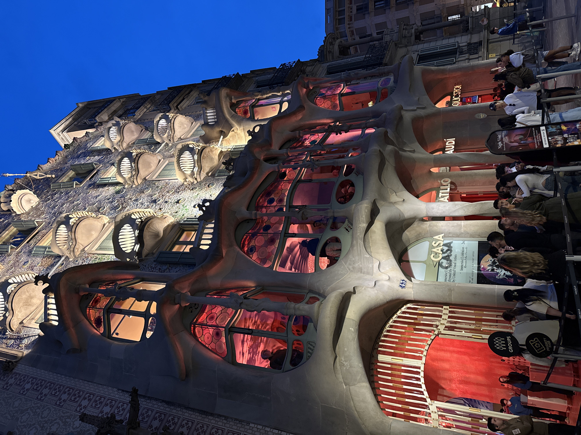 The image captures the illuminated façade of Casa Batlló, a renowned architectural landmark in Barcelona, Spain, designed by Antoni Gaudí. A crowd of people, some standing in line, is visible in front of the building's entrance, indicating it is likely a popular tourist destination. Signage displays information about tours and exhibits, including brands like "Audi" and "GROUPS PRIORITY PASS," suggesting sponsored or tiered access. The distinctive organic shapes and colorful windows of the building are prominently featured, as well as the blue sky on the upper left corner.