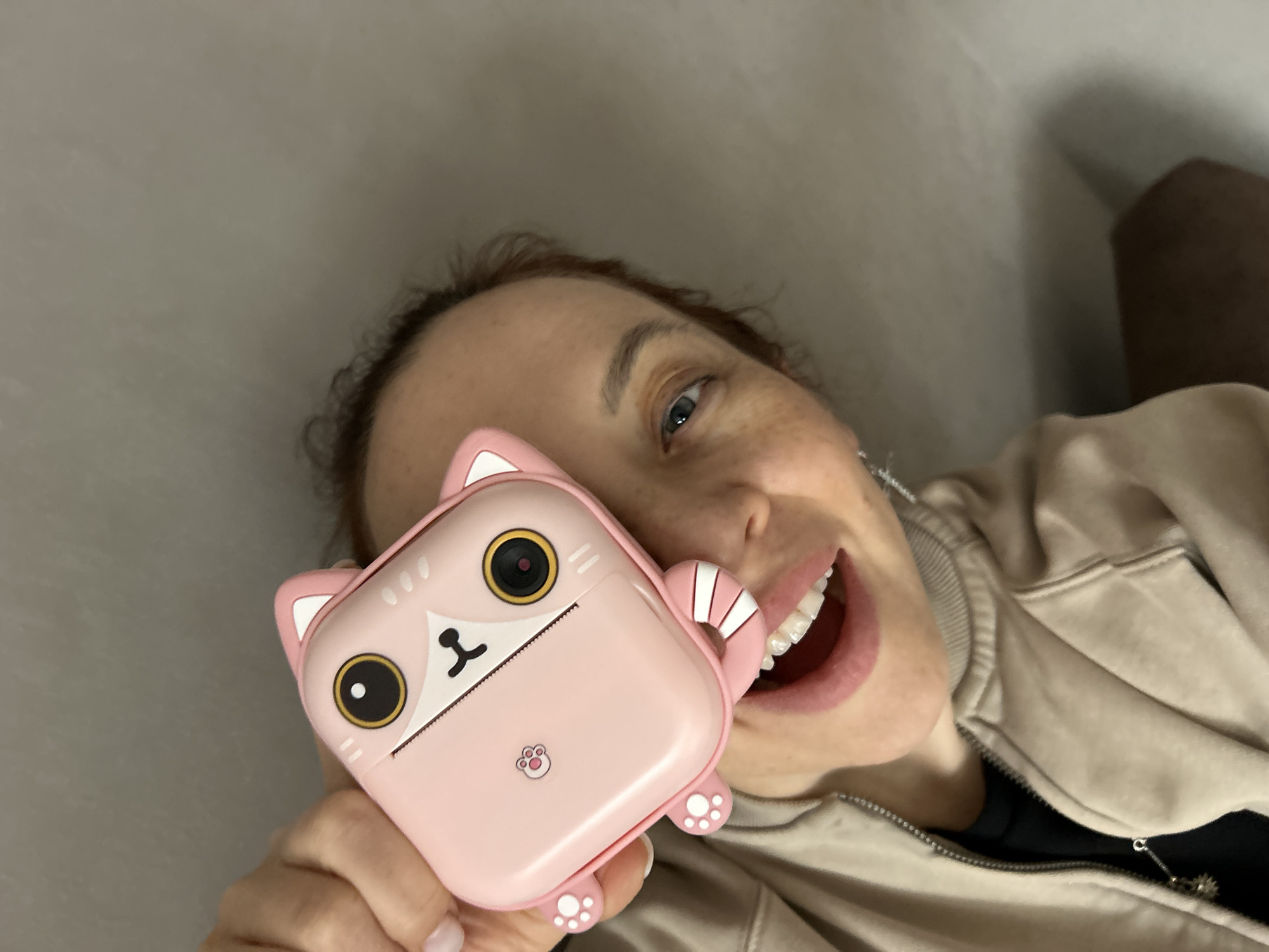A woman is holding a pink, cartoon-cat-shaped object up to her face. The object has large, round, black and white eyes, a small black nose and mouth, and tiny white paws on the bottom. The woman is smiling broadly, showing her teeth. She has fair skin, red hair, and freckles. She's wearing a light brown jacket and a black shirt. The background is a plain, light gray wall. It's not possible to verify if the photo was taken in Chaikovsky, Russia, based on the visual information available.