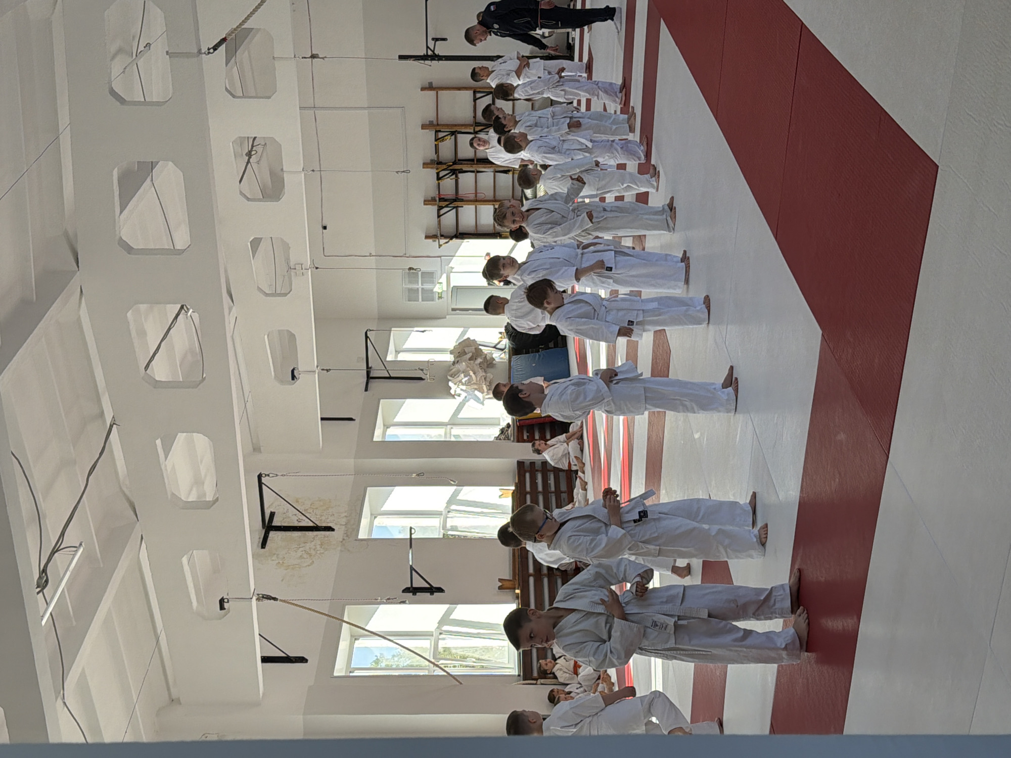 A group of children in white martial arts uniforms, presumably judo gis, are standing in a straight line in a training hall. They appear to be barefoot. A man in a dark suit is standing at the head of the line. The room has a white ceiling with structural supports and windows along one wall. The floor is white with a wide stripe of red matting running parallel to the line of children. The location is likely a martial arts dojo in Chaikovsky, Russia.