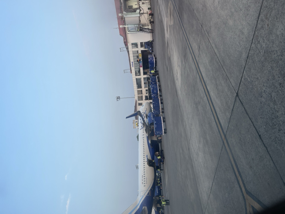 The image, taken in what appears to be daytime under a clear, light blue sky, depicts an airport tarmac scene, likely at Guwahati, India. A large Indigo Airlines aircraft, predominantly white with blue livery and tail, occupies the left side of the frame. Its registration number, VT-INR, is visible near the front underbelly.
Various pieces of ground support equipment are positioned around the aircraft, indicating active servicing. These include multiple blue luggage carts or cargo loaders connected in a train, and mobile stairs leading up to the plane's door. Several ground crew members, wearing high-visibility vests, are visible near the plane and the equipment, engaged in tasks such as loading or unloading.
To the right of the aircraft, an airport terminal building stretches horizontally across the midground. It is a multi-story structure with a light-colored facade and a reddish roof. The word "Arrivals" is clearly legible on one section of the building's exterior. Jet bridges are connected to the terminal, and some ground vehicles are parked near the building.
The tarmac in the foreground and midground is a grey concrete surface with yellow and dark grey markings, indicating pathways and parking areas. The overall impression is one of a busy but orderly airport operation under bright, fair weather conditions.
