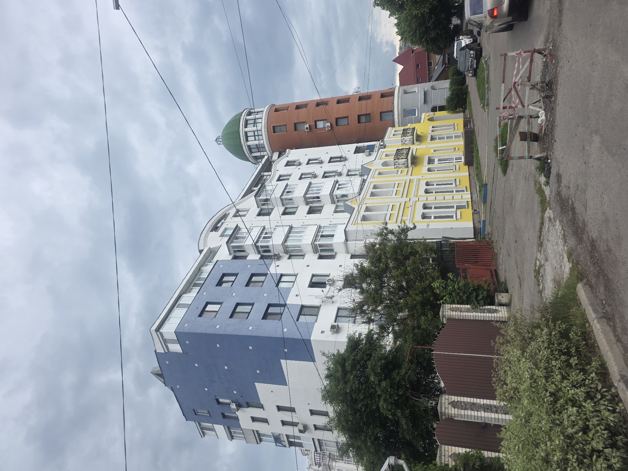 The image presents an urban street scene in Bila Tserkva, Ukraine, under an overcast sky. The focal point is a cluster of buildings exhibiting diverse architectural styles.
On the left, a multi-story residential building dominates, characterized by a contemporary design. Its facade is predominantly white, featuring large, irregular sections of muted blue. Numerous rectangular windows and external air conditioning units are visible across its surfaces. Green deciduous trees partially obscure the lower levels of this building.
In the center and midground, a two-story building with a bright yellow facade and white trim stands out. It features multiple tall, arched windows and decorative black wrought-iron balconies. Immediately to its right is a smaller, modern-style building with a curved grey facade. Further right, a prominent, tall, cylindrical tower with a reddish-brown brick exterior rises, crowned by a distinct green dome. This tower also features multiple rectangular windows and external air conditioning units. Behind the grey building and to the right of the tower, a structure with a steep red roof is partially visible.
The foreground consists of an unpaved, uneven dirt and gravel area that transitions into a paved road on the right. A concrete curb separates the dirt path from a dense patch of green and white variegated shrubs. In the immediate foreground, a dilapidated, makeshift wooden barrier, adorned with red and white caution tape, lies on the ground, indicating a potential obstruction or area of work.
On the paved road to the right, several parked vehicles are visible, including a dark sedan (possibly a Mercedes-Benz) and a lighter-colored sedan.
The sky is uniformly covered with thick, grey clouds, suggesting an overcast day. Multiple dark electrical or utility wires stretch horizontally across the upper portion of the frame. No individuals are present in the visible scene.
This specific location is identifiable as Yaroslava Mudroho Street in Bila Tserkva, Ukraine, featuring the distinctive landmark known locally as the "House with a Dome" or "House with a Tower" (Будинок з куполом / Будинок з вежею).