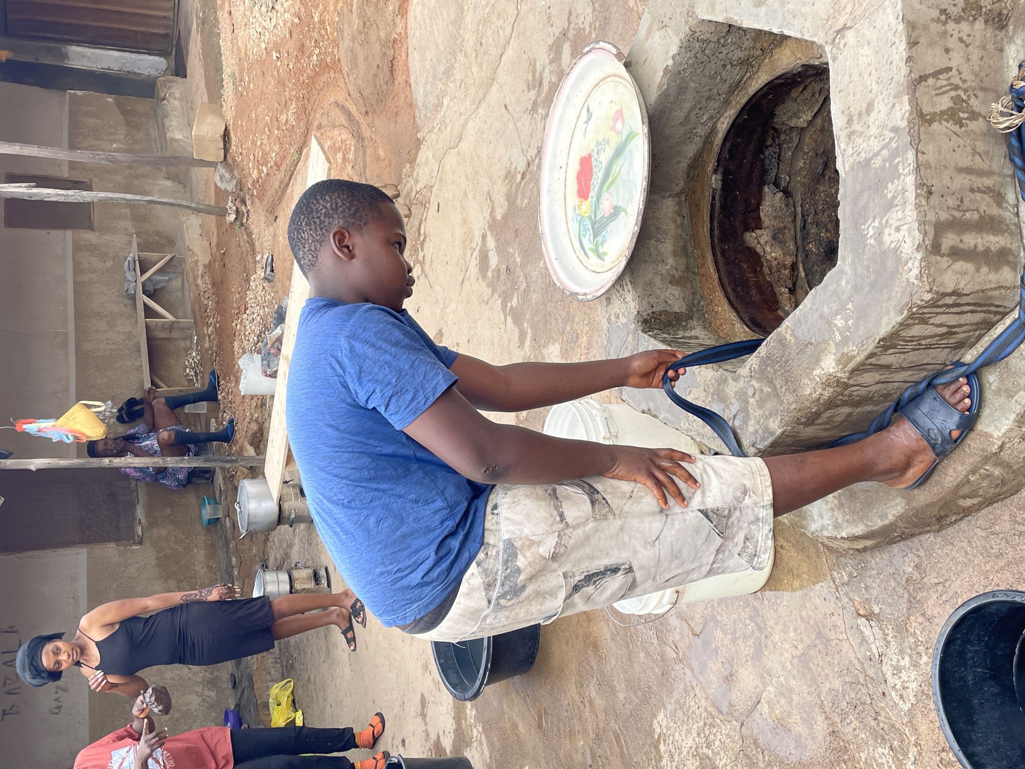 In Jos, Nigeria, a young boy wearing a blue t-shirt and camouflage shorts is seated near a stone well, with a decorative metal lid leaning against it. He holds a blue rope, presumably attached to a bucket inside the well. Several white plastic buckets are nearby, along with a large black bucket on the ground. In the background, two women are visible, one in a black dress and another in an orange shirt, while another individual is lying on a platform. The setting appears to be a concrete courtyard or common area, adjacent to a building structure.
