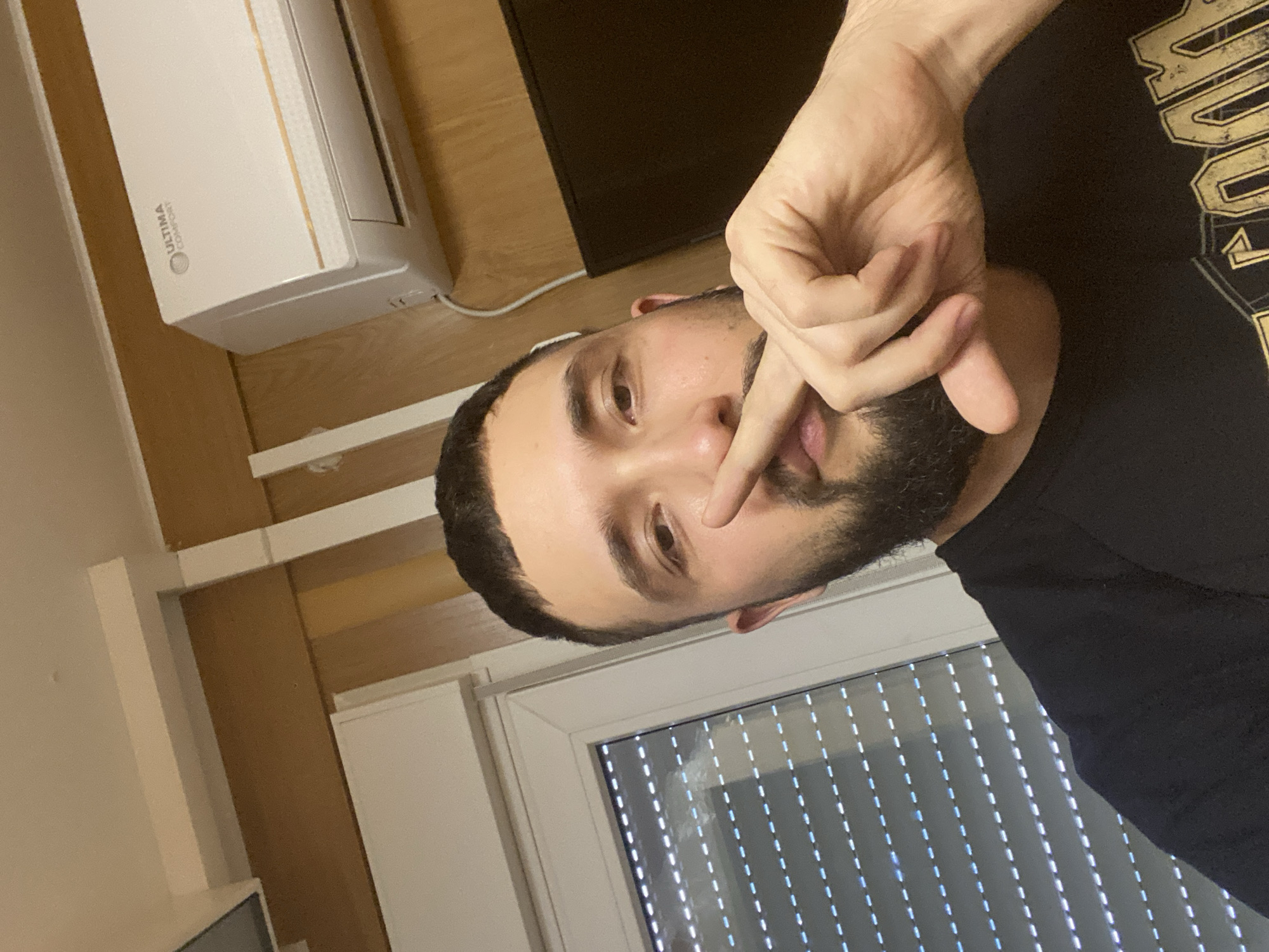 A man with dark hair and a beard is holding his finger to his lips. He is wearing a black t-shirt with a gold design. Behind him is a window with vertical blinds. To the left of the man is a white air conditioning unit mounted on the wall. The wall appears to be wood paneling. The location is implied to be Koskolovo, Russia, but the image does not provide specific geographical or contextual information to verify this.