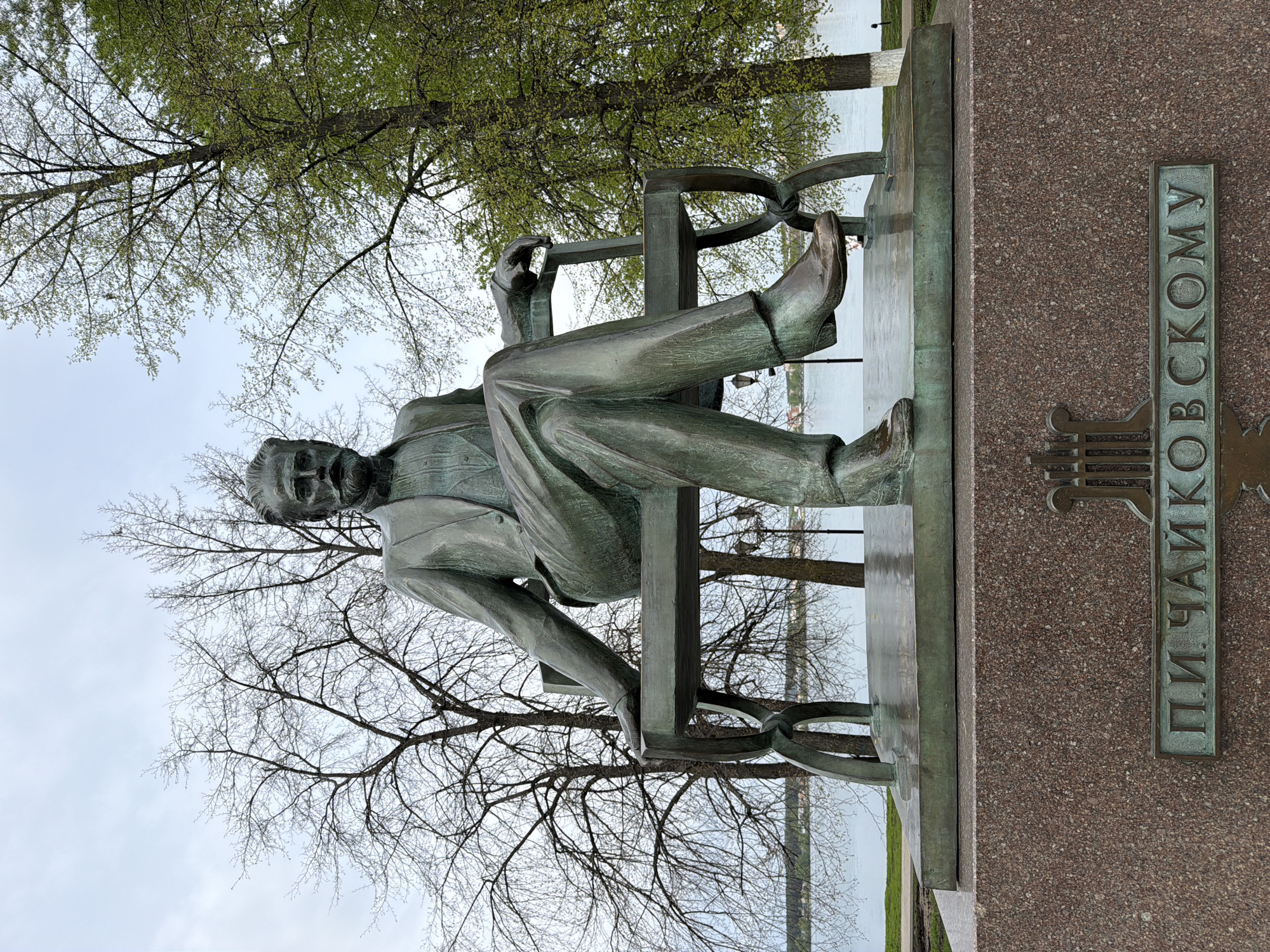 The image depicts a bronze statue of Pyotr Ilyich Tchaikovsky in Votkinsk, Russia. The statue shows Tchaikovsky seated in a chair, with his legs crossed. The base of the statue includes a plaque with his name written in Russian. The statue is outdoors, with trees in the background and what appears to be a body of water behind the trees. The sky is overcast.