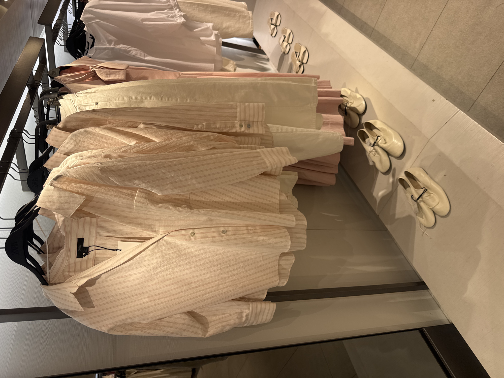 A close-up, eye-level shot shows a rack of women's clothing in a retail store, possibly in Kuta, Indonesia. The garments, primarily in pastel shades of peach and cream, are neatly hung on black hangers. The foremost item is a light peach, striped button-down shirt with a relaxed fit and a pointed collar. Behind it, several other garments, including dresses and blouses, are visible. On the floor below the clothing rack, a pair of cream-colored flat shoes are displayed, and further to the right, a row of three pairs of cream-colored slip-on sandals are arranged. The background consists of a wall with a muted, textured finish and a large mirror on the left side of the frame, reflecting a portion of the store. The lighting appears to be typical retail store illumination. There are no people visible in the image.
