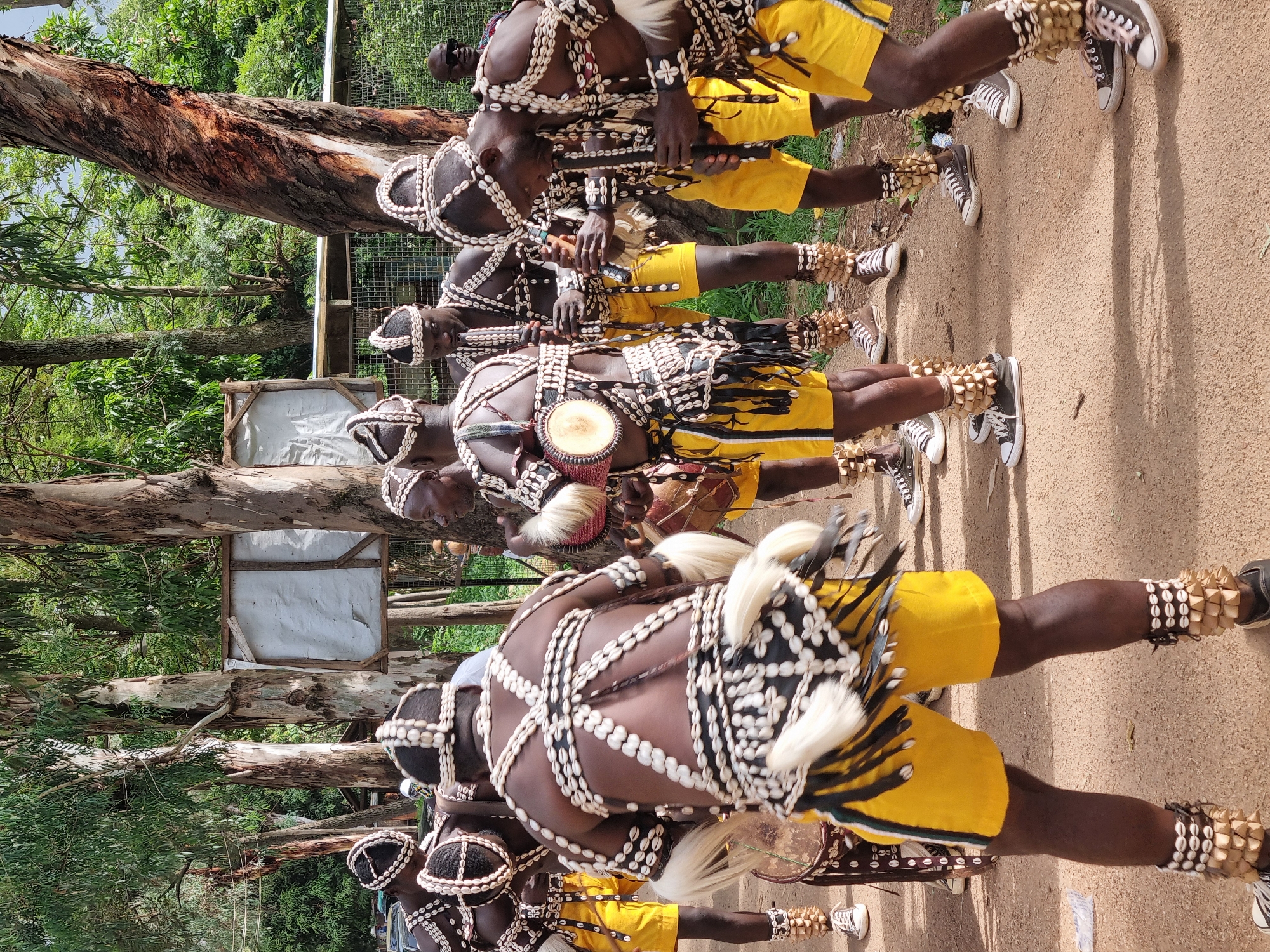 A group of men in traditional attire are gathered outdoors, likely in the city of Jos, Nigeria. The men are dressed in yellow shorts and ornate tops adorned with shells, beads, and white fringe. Some of them are holding and playing musical instruments, including what appear to be flutes and drums. The background shows tall trees and some rudimentary structures, suggesting a rural or semi-rural setting within the city. The ground is dirt. The lighting indicates it is daytime, likely sunny, with dappled shade from the trees. The men appear to be participating in a cultural performance or ceremony.