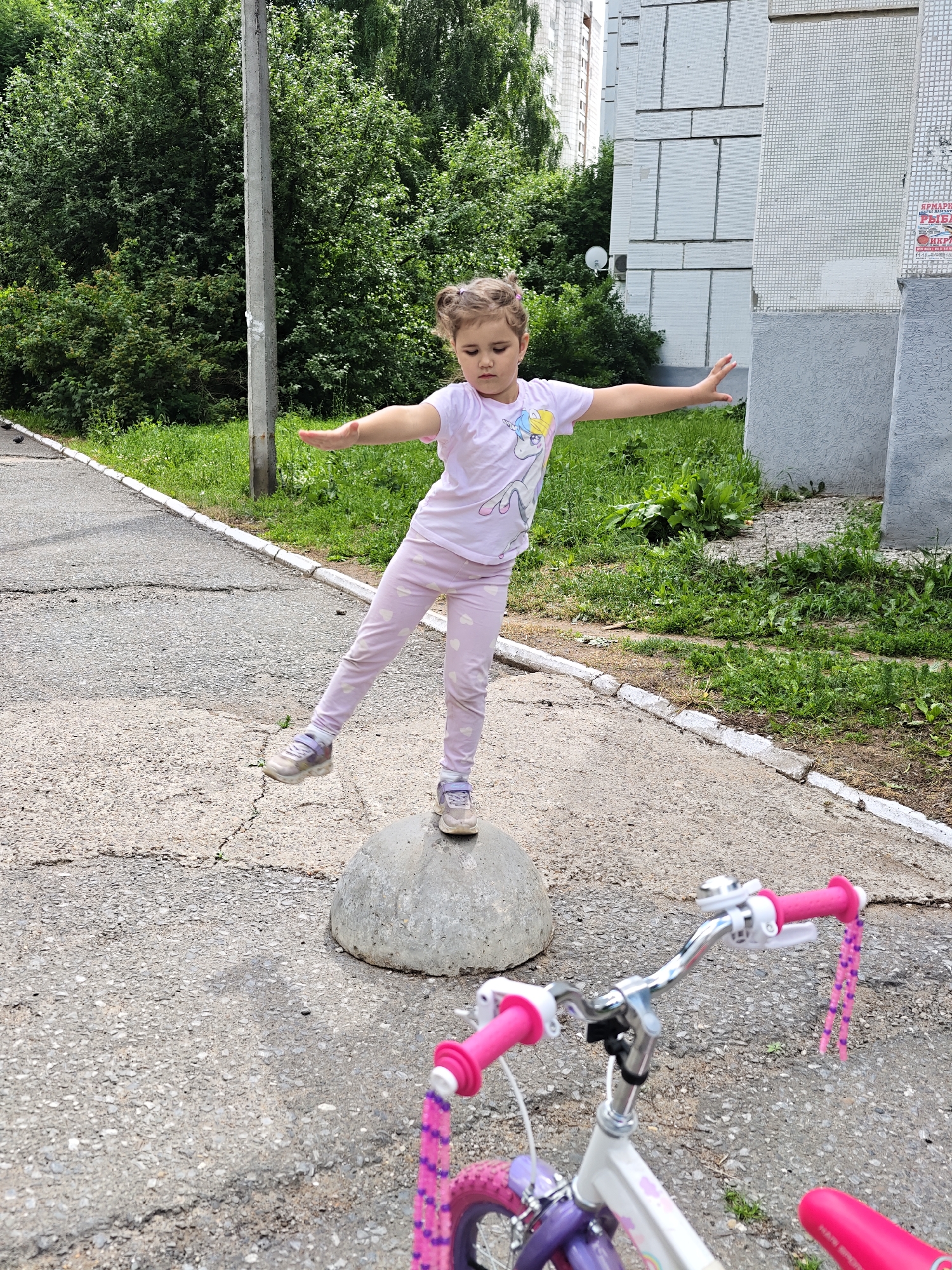 A young girl, appearing to be around 5 years old, stands on a large, rounded concrete bollard on a paved surface. She wears a light pink t-shirt with a unicorn graphic and light pink leggings with a heart pattern. Her arms are outstretched horizontally, and her right leg is extended backward, suggesting she is balancing or posing. She has her hair pulled up in two small buns. In the foreground, a portion of a white and purple bicycle with bright pink handlebars and pink streamers is visible. The background consists of lush green trees and a light-colored, multi-story building with a tiled facade, characteristic of urban residential architecture. The location is identified as Chaikovsky, Russia. The overall scene depicts an outdoor recreational activity in a semi-urban environment.