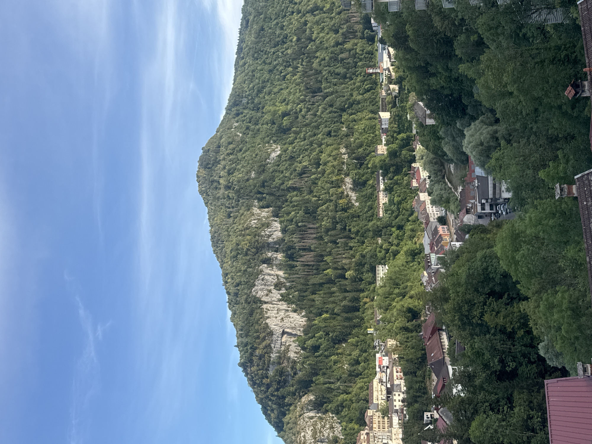The image is a scenic view of a town nestled at the base of a large, tree-covered mountain. The sky is a clear, bright blue with faint wispy clouds. The mountain dominates the upper half of the image, with its slopes covered in dense green foliage, interspersed with rocky outcrops and patches of lighter-colored rock. The lower portion of the image reveals a town with buildings of various sizes and architectural styles, mostly in muted tones of beige, white, and red. Some buildings appear to be residential, with tiled roofs, while others seem to be commercial or public structures. Trees are abundant throughout the town, creating a harmonious blend of nature and urban development. The presence of parked cars on a street suggests the town is inhabited. The foreground features the tops of some red-tiled roofs, indicating the vantage point from which the photograph was taken. The overall impression is one of a peaceful, picturesque town surrounded by natural beauty, likely Saint-Claude, France, given the context.