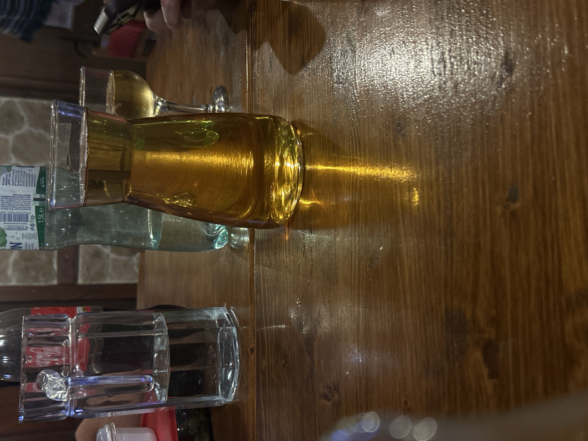 A high-angle shot depicts a wooden table setting with assorted beverages and glassware. A full carafe containing a golden liquid sits next to a wine glass filled with a similar colored drink. A water bottle and other glasses are also present on the table. Part of a hand holding a dark object is visible. The table reflects light, revealing its grain. The location is Mirovyane, Bulgaria.