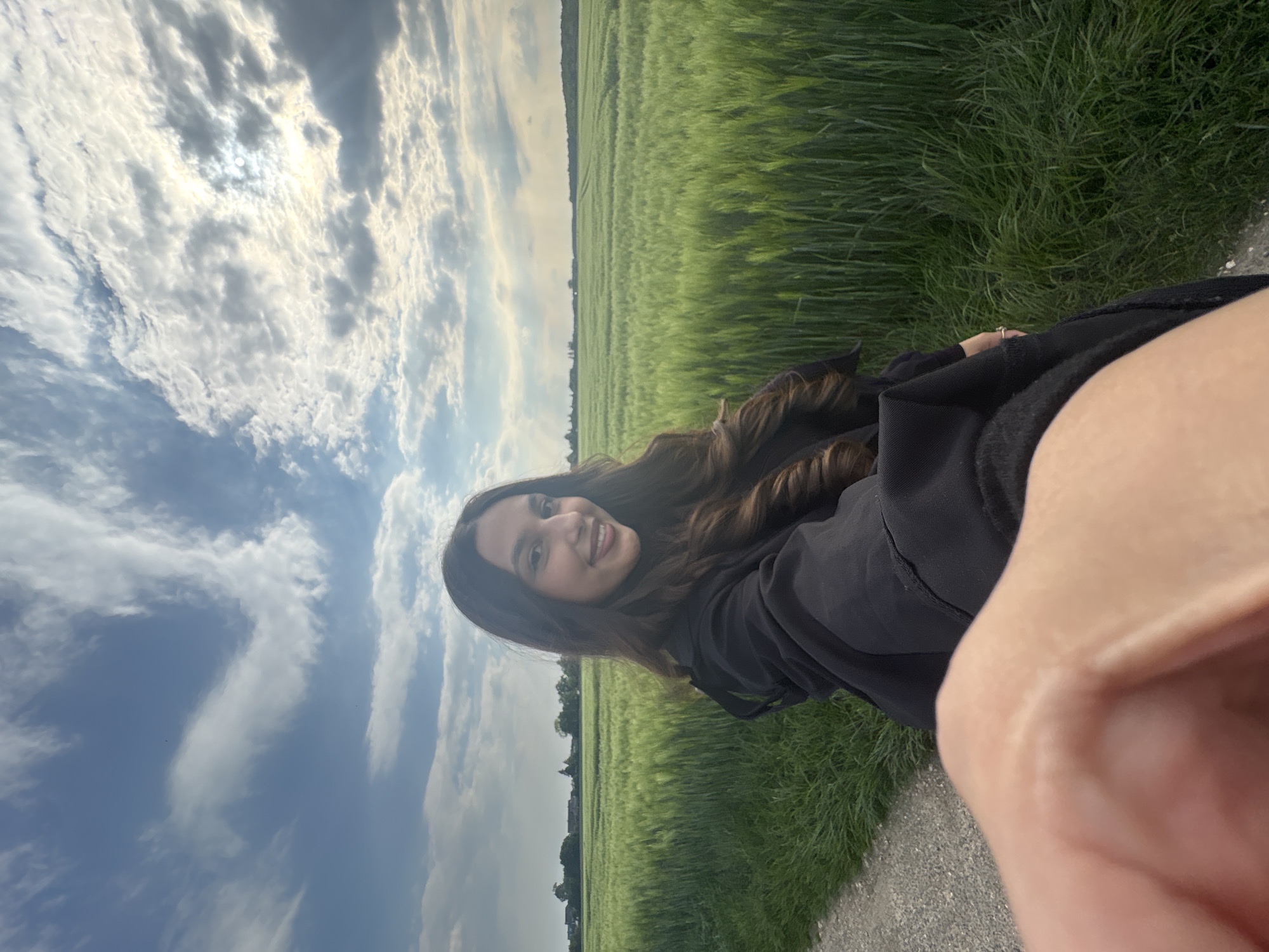 A selfie photo of a young woman with long brown hair, smiling at the camera. She wears a black hooded jacket. She stands on a gray paved road, with a field of tall green grass behind her. The sky above is filled with puffy white clouds. Based on the location details, the scene is likely in or near Wegberg, Germany. The foreground is partially obscured by what appears to be the photographer's hand holding the camera.