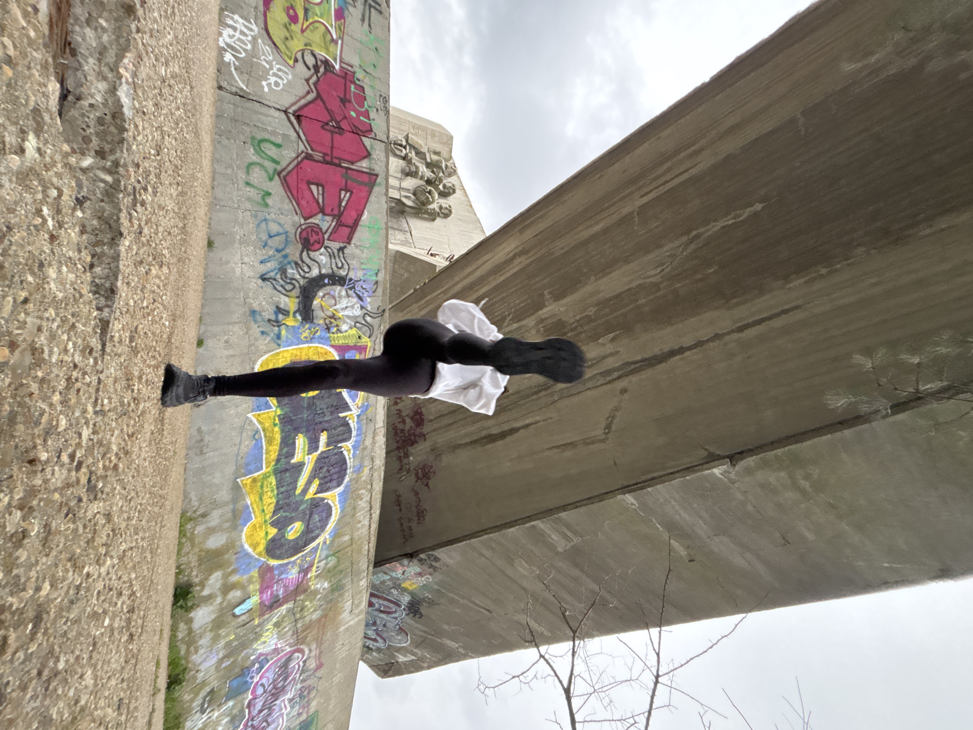A low-angle, rotated photograph captures a person posed in an apparent handstand position against a graffitied concrete bridge support. The individual is dressed in black leggings and a white top, their feet pointed towards the left edge of the frame. The graffiti on the support features a variety of colors and styles, including block lettering, cartoonish figures, and abstract designs. The bridge structure itself is made of concrete and appears aged, with visible wear and staining. In the background, additional bridge supports are visible, along with a cloudy sky. Bare tree branches are visible in the lower right corner of the image, suggesting a time of year with sparse foliage.