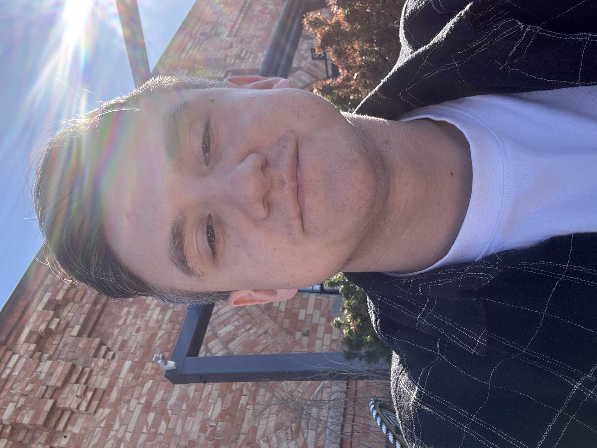 A man is photographed in an outdoor setting. He has short hair and is wearing a white t-shirt under a dark plaid jacket. The background includes a brick wall and some foliage. The sun is bright and casting a lens flare in the upper left corner of the image. The subject is facing the camera with a slight smile. The location is unknown and cannot be assumed to be Warsaw, Poland without additional information.