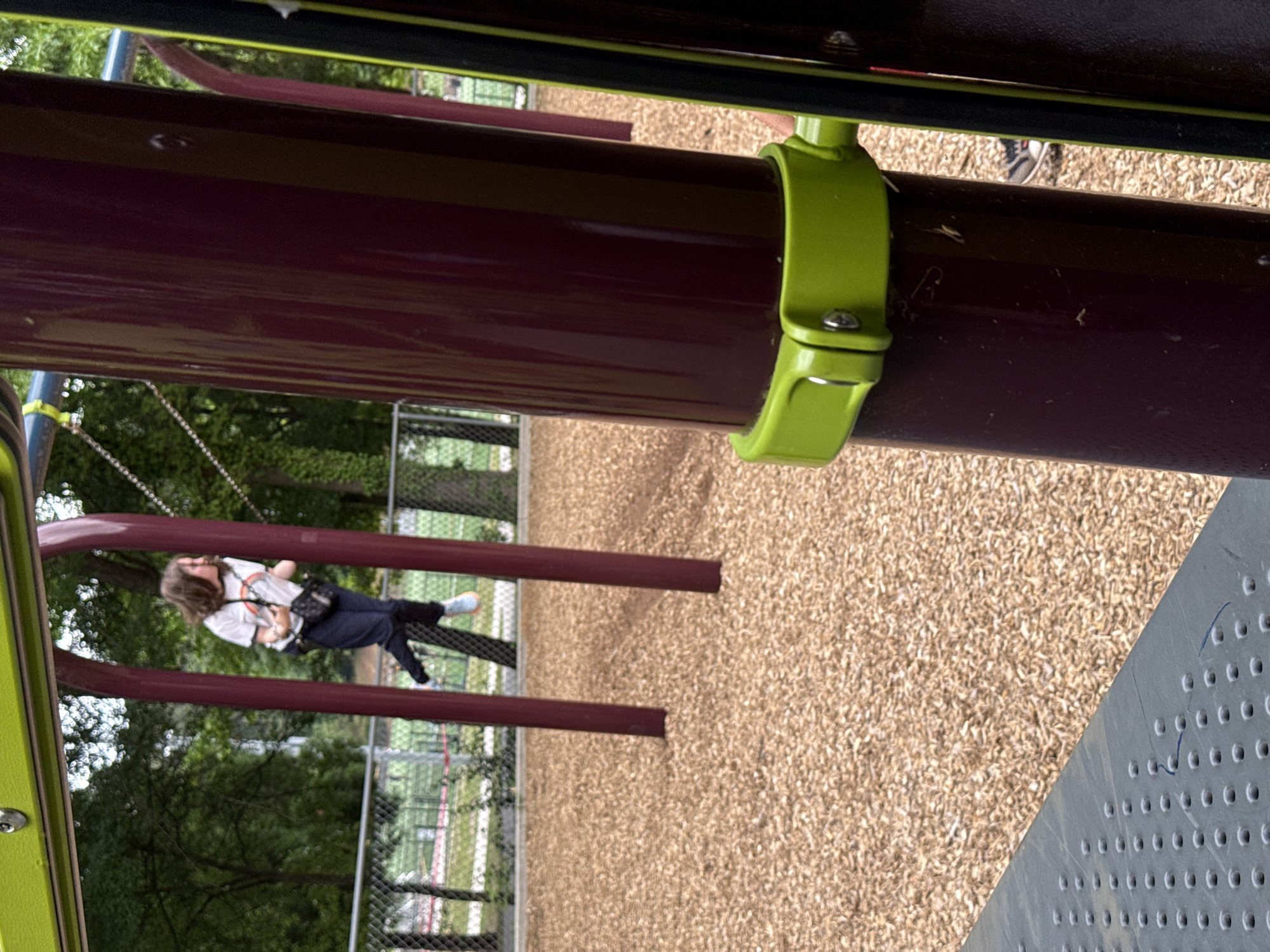 The image shows a playground in Mountain Park, USA. A girl is swinging on a swing set. The playground surface appears to be covered with wood chips. The equipment consists of maroon-colored metal bars and green support clamps. A perforated gray metal platform is visible in the foreground. A chain link fence encloses the area. The environment suggests an outdoor recreational space within a residential or park setting.