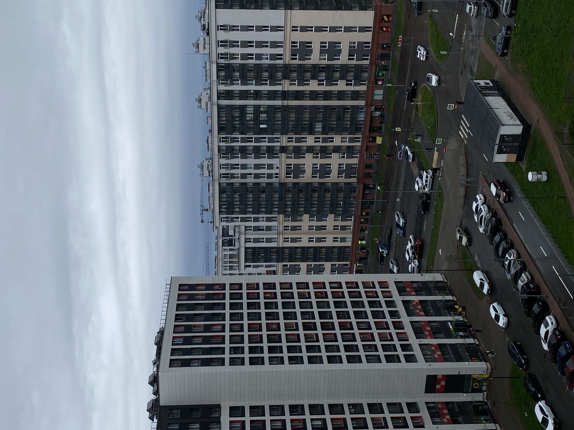The image shows a view from an elevated position, likely a high-rise building, in an urban area, suspected to be Unknown, Russia based on visual cues. The sky is overcast. The scene includes two modern apartment buildings with a grid-like facade of windows and a color scheme of light gray, dark gray, and red details. The building in the foreground has a prominent white exterior with red accents around the windows. A street-level view includes multiple parked cars, a bus stop-like structure, pedestrian crosswalks, and intersections. The vehicles are a mix of colors, predominantly white and dark-colored sedans and SUVs. A small green area with grass is visible near the road. A construction crane is visible in the distance between the buildings. A pedestrian is visible at the zebra crosswalk.