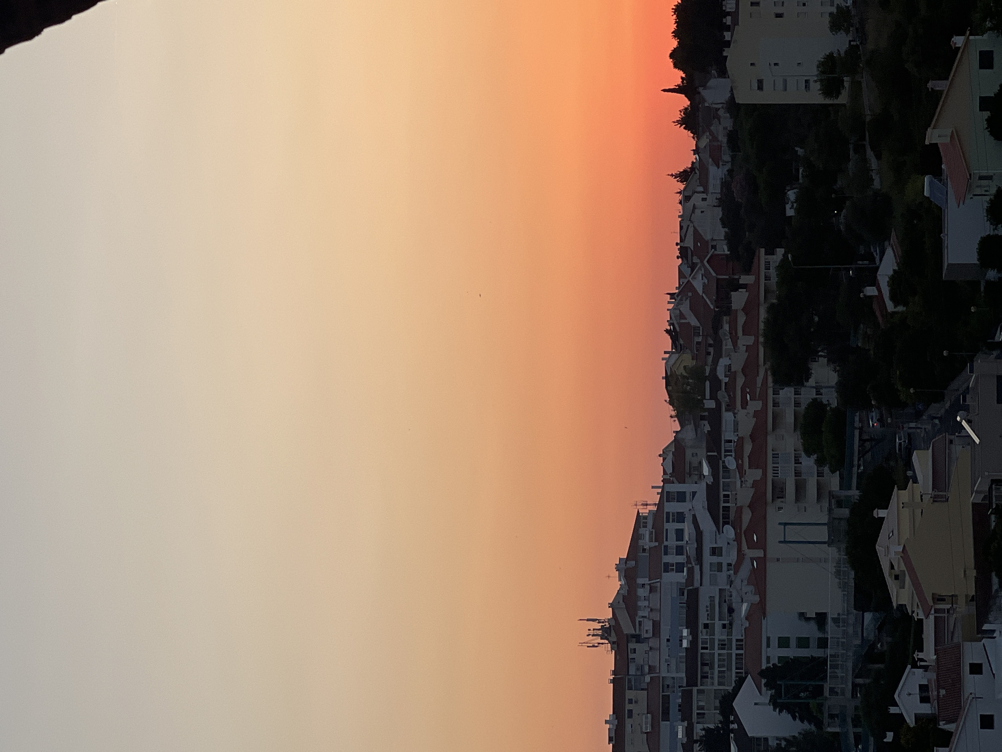 The image depicts a view of Cascais, Portugal, at sunset. The sky transitions from a pale yellow at the top to a vibrant orange and pink near the horizon. A dense residential area with buildings of various sizes and architectural styles occupies the midground and foreground. Many buildings have red-tiled roofs. Some buildings are taller, multi-story structures, while others are smaller houses. Trees and dark foliage are interspersed among the buildings, especially in the lower part of the image. A few cell towers are visible on top of one of the buildings. The scene is quiet, with no people or vehicles actively engaged in any discernible actions. The overall impression is of a residential neighborhood viewed from an elevated position during twilight.