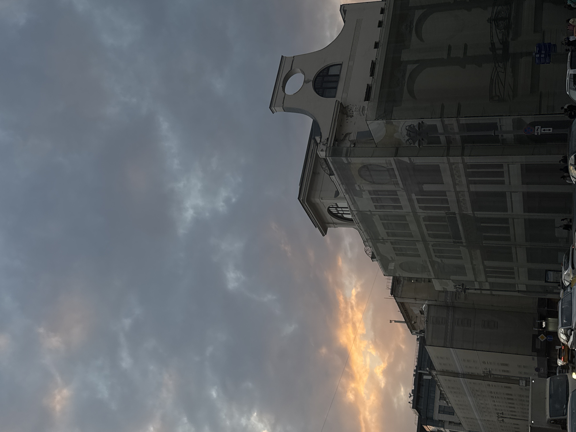 The image shows a street scene in Moscow, Russia, at dusk. The sky is filled with dramatic, textured clouds, with a warm glow of sunset breaking through in the middle right of the frame. Buildings line the street, some with detailed architectural features and painted murals or embellishments. One building in the foreground has a distinct curved facade with a circular opening. Cars are visible parked along the street, some with their headlights on, indicating the fading light. A few people are discernible in the scene, walking on the sidewalk, adding a sense of daily life to the urban setting. The overall mood is atmospheric and somewhat melancholic due to the overcast sky and the low light. The image is oriented vertically, with the sky taking up a significant portion of the frame. No discernible text is visible.