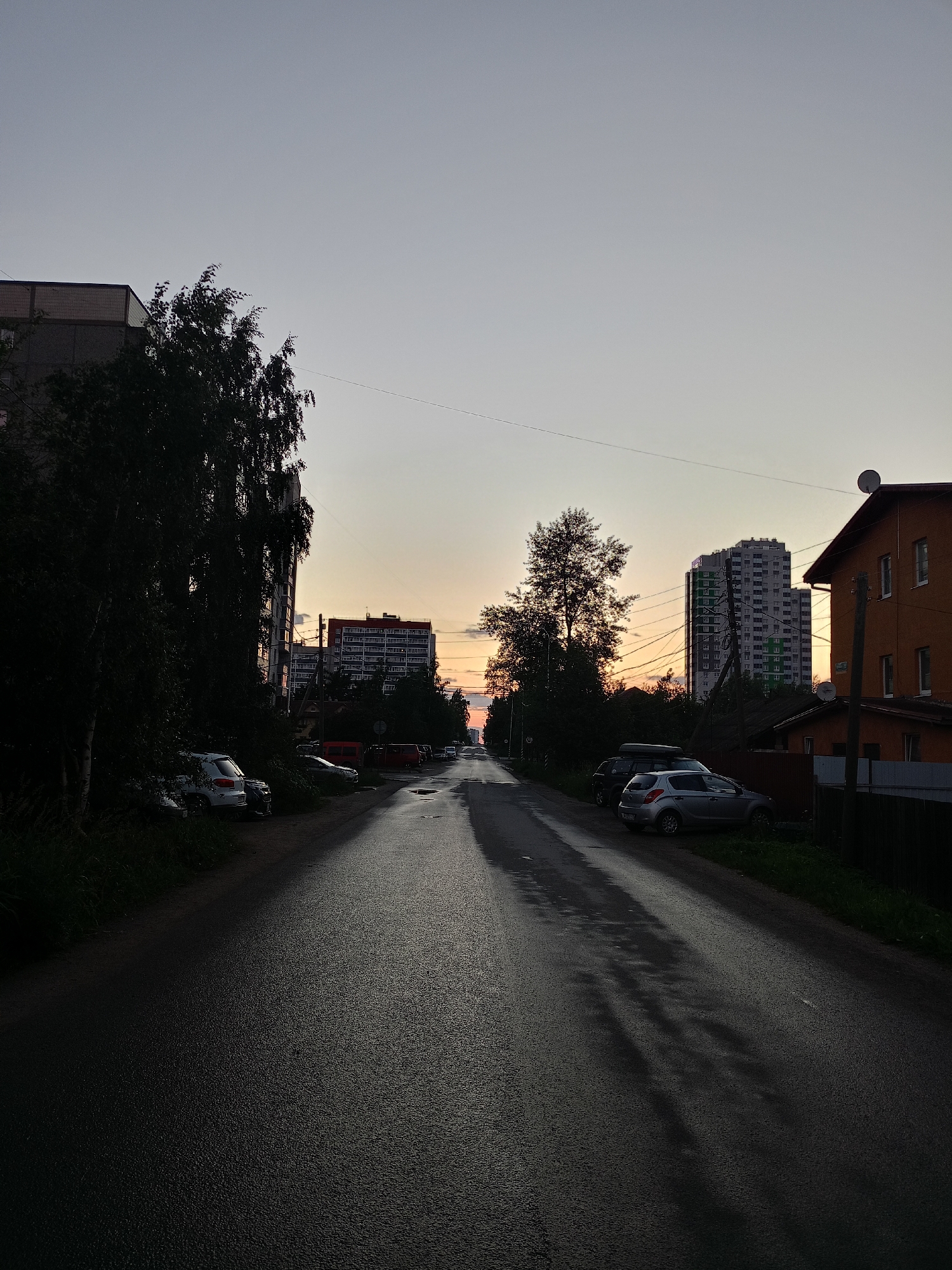 A street scene at dusk in an urban environment. The sky is a gradient of pale gray at the top transitioning to pale orange near the horizon. A wet asphalt road stretches into the distance, reflecting the dim light. On the left side of the road, a tall, multi-story apartment building is partially visible behind silhouetted trees. Parked cars are visible along the side of the road. On the right side of the road, a low-rise, orange building with a dark roof and windows is present, along with a wooden fence. Further down the road, more multi-story buildings are visible in the distance, illuminated by the fading light. A few more cars are parked along the right side of the road. The scene is devoid of any people.