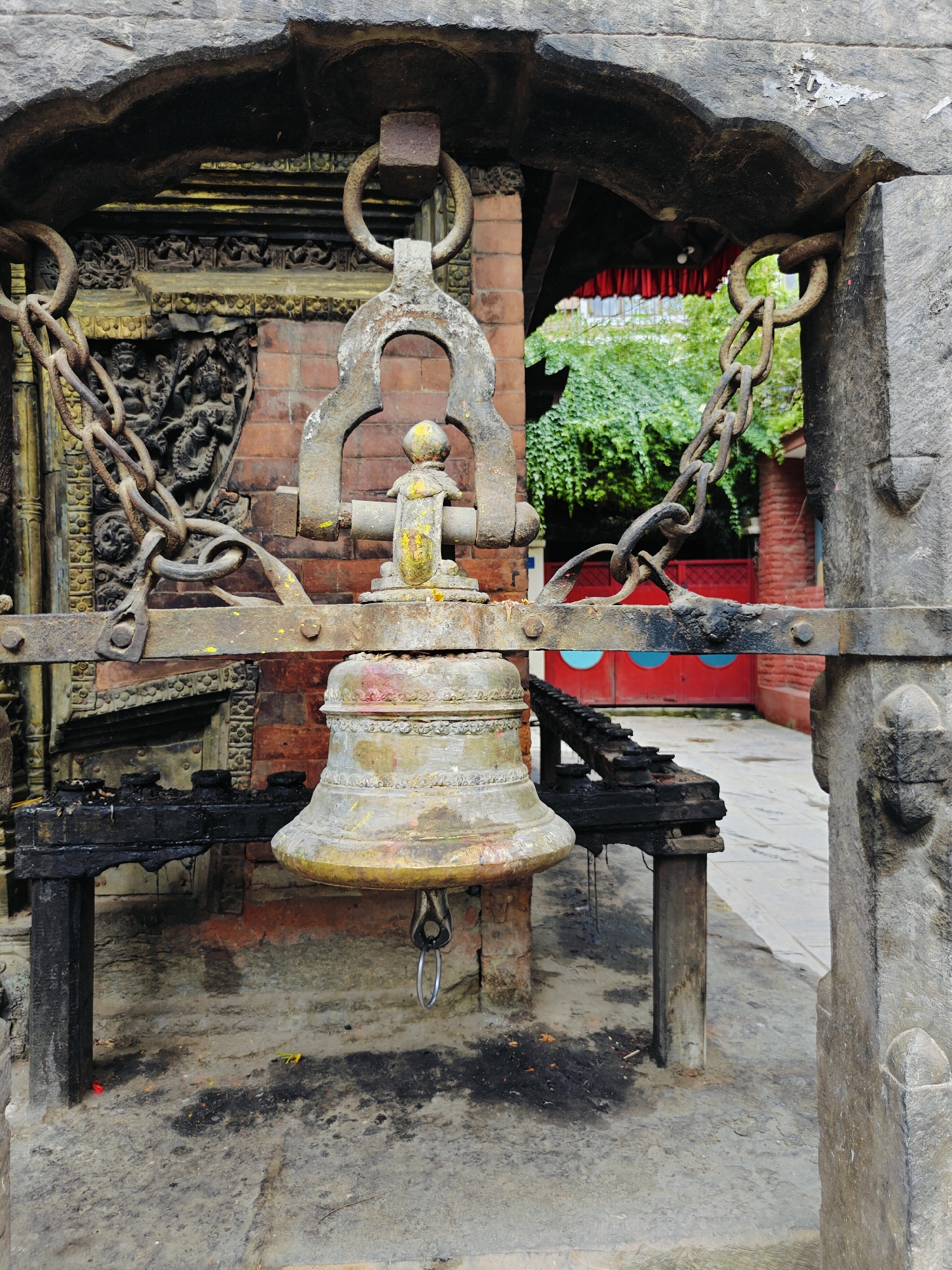 This is a medium shot, taken at eye-level, of a large, ornate bell hanging from chains within an ancient, stone structure. The bell, made of aged metal, appears to be weathered and possibly painted with hints of red and yellow pigment. It is suspended from a crossbar, which is itself supported by thick, rusty chains. The chains are attached to large, weathered stone pillars that frame the scene.
In the background, a red building with arched windows is visible, partially obscured by lush green foliage. A red curtain hangs above an opening in the structure. The ground is paved with stone and appears somewhat damp, with dark patches and scattered debris, suggesting it is an outdoor, public space. The overall impression is one of age and historical significance. This scene is likely from the city of Lalitpur, Nepal, a place renowned for its ancient temples and rich cultural heritage.