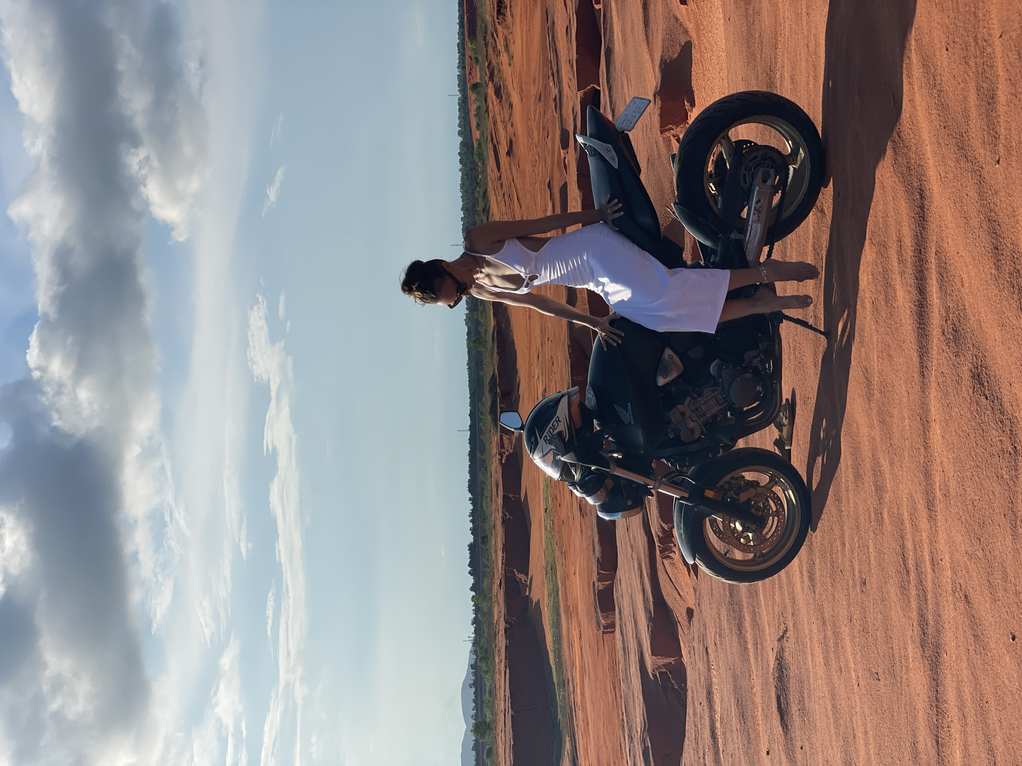 A woman in a white dress sits on a black motorcycle in the desert landscape of Mui Ne, Vietnam. The woman is positioned facing left, with her legs crossed and her arms resting on the motorcycle seat. She wears sunglasses and her hair is pulled back in a bun. The motorcycle is a Honda, with a helmet resting on its handlebars. The desert features red sand dunes and sparse vegetation in the background, with a partly cloudy sky above.