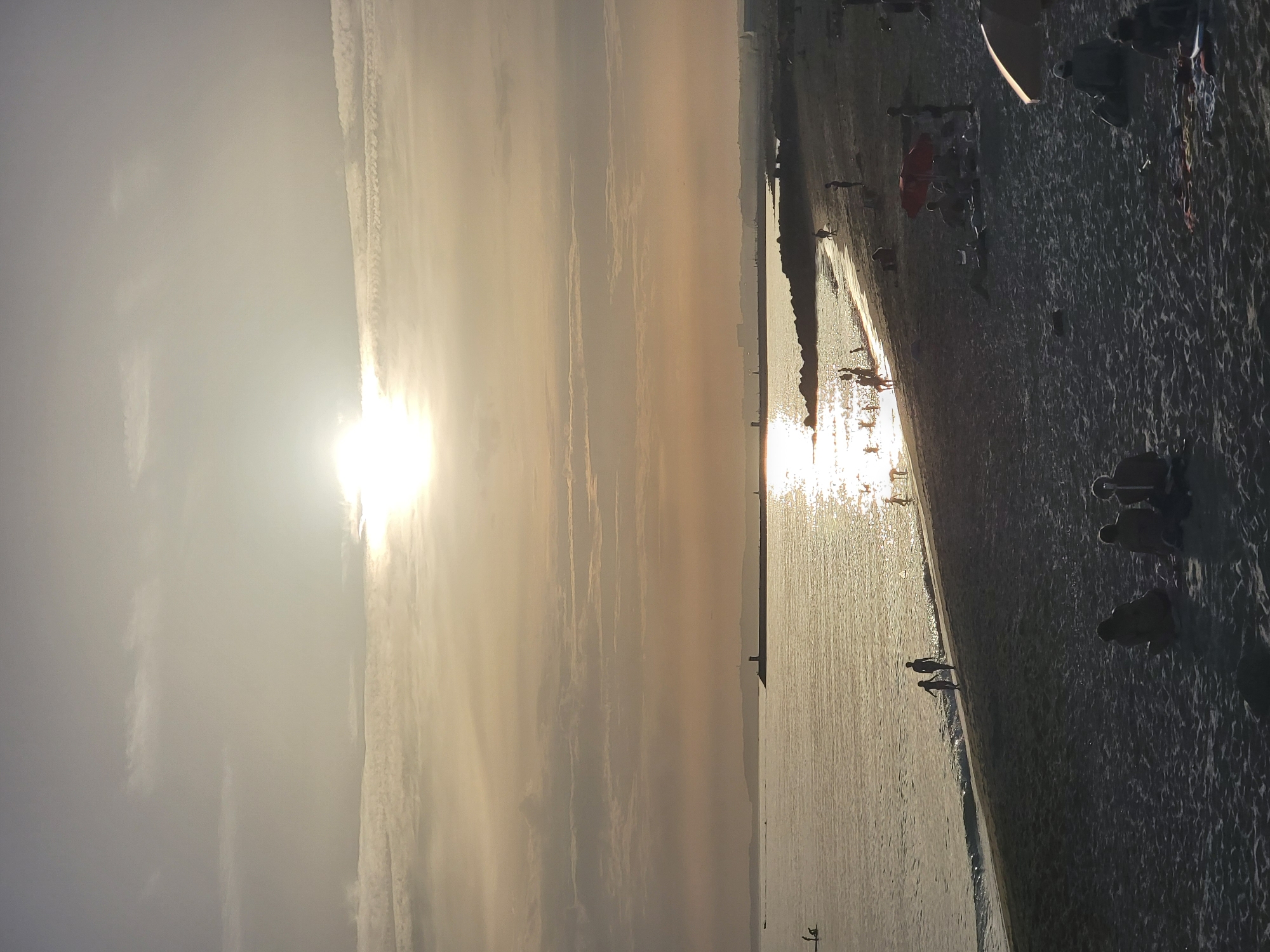 This is an outdoor, eye-level shot of the Quarteira, Portugal coastline during sunset. The sun, a bright white orb, is positioned slightly right of the center of the frame, low in the sky and partially obscured by thin, wispy clouds. Its reflection creates a shimmering path across the calm, golden-hued ocean water. The water meets a sandy beach lined with dark rocks that form a breakwater or jetty.
Scattered across the beach and shallow water are several individuals and groups of people. Some are wading or playing in the water, while others are walking along the shoreline. Closer to the foreground, groups of people are seated on the sand, some under umbrellas. A few buildings and a lighthouse are visible in the distant hazy background, along the coastline. The overall atmosphere is serene, with the warm tones of the sunset casting a soft light over the scene.