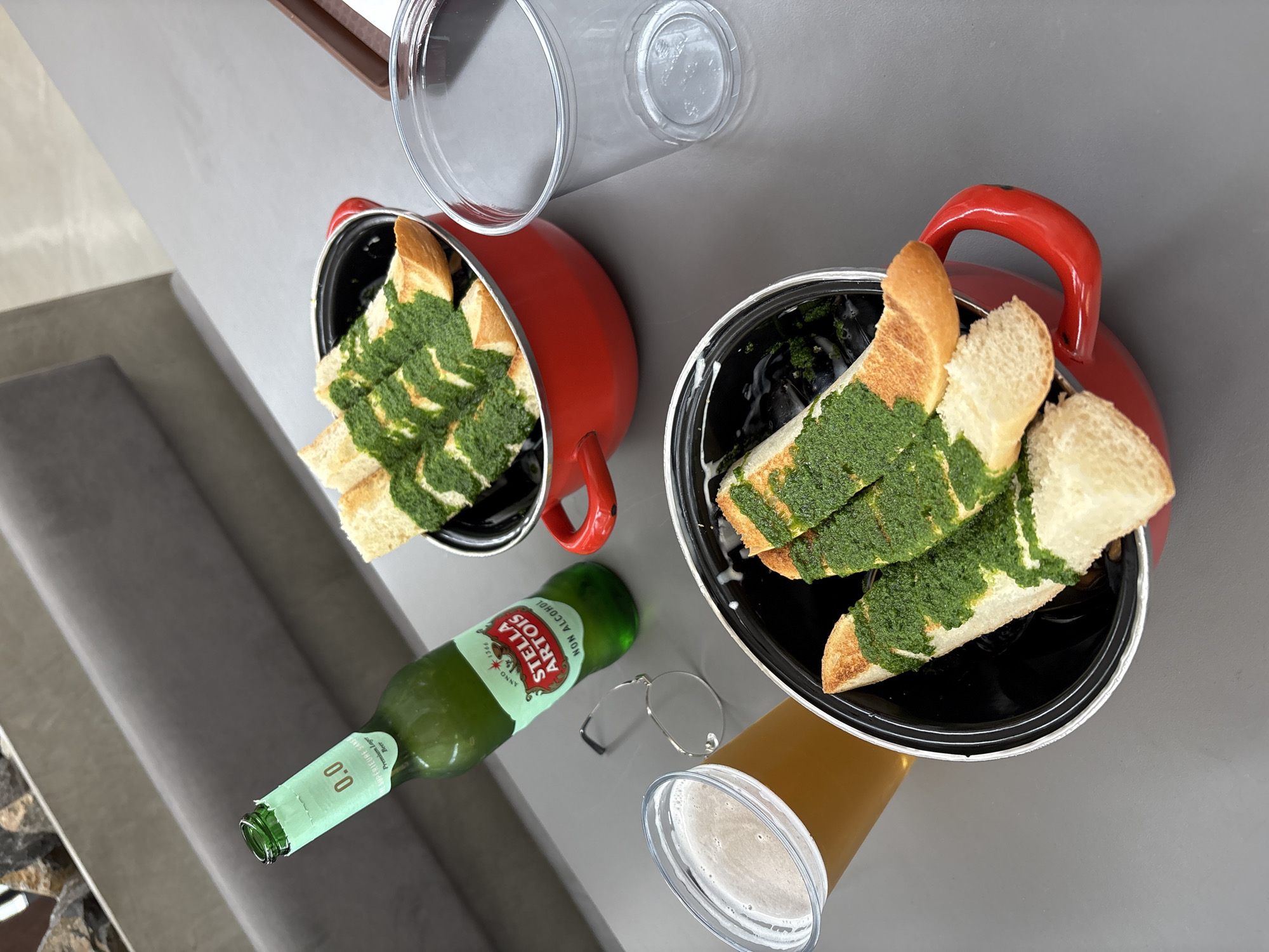 The image depicts a table setting with two small pots of bread topped with a green sauce, likely pesto, positioned diagonally across the frame. A bottle of Stella Artois 0.0% non-alcoholic beer lies on its side between the pots. Next to the beer is a pair of eyeglasses and a clear plastic cup filled with a brown liquid, possibly beer or tea. Two additional clear plastic cups are also present. The background is a neutral gray surface, with a glimpse of a gray upholstered bench. Based on the presence of Stella Artois and the style of the food presentation, the location is likely a restaurant or cafe. The city of Krasnoyarsk, Russia, cannot be determined from the image itself.