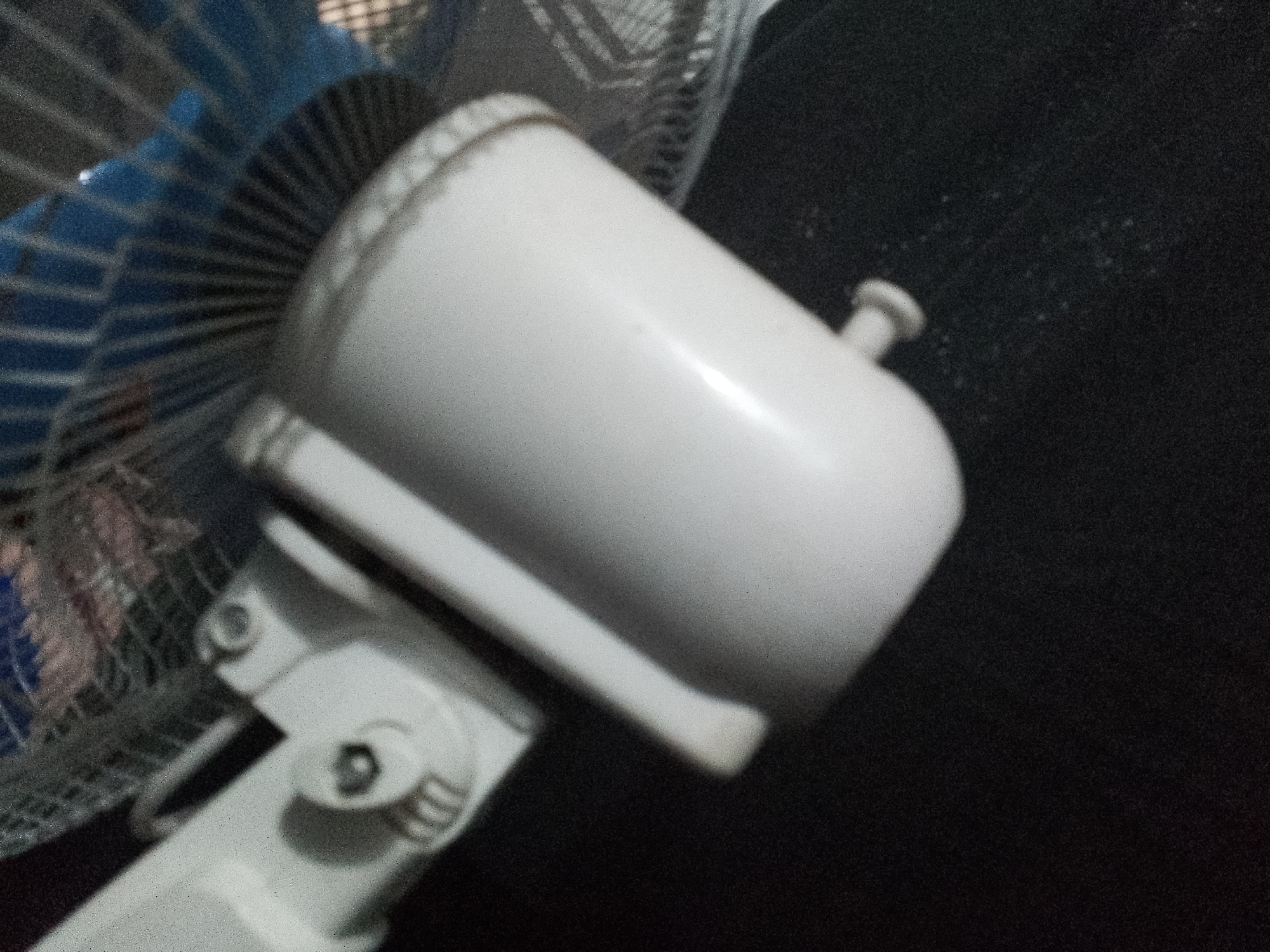 The image shows a close-up, angled view of the motor housing of a white electric fan. The fan's blue grill is visible on the left side of the frame, partially obscuring the fan blades. The motor housing is cylindrical with a textured top edge and a small, protruding control knob. A white support arm extends from the bottom of the housing, connecting to a more complex joint with visible screws and adjustment mechanisms. The background is a dark, textured surface, likely fabric, with some dust or debris visible. The lighting is somewhat harsh, creating a bright highlight on the motor housing. The scene is a static, inanimate object in an indoor setting, with no people or animals present. The location is specified as Unknown, Unknown.
