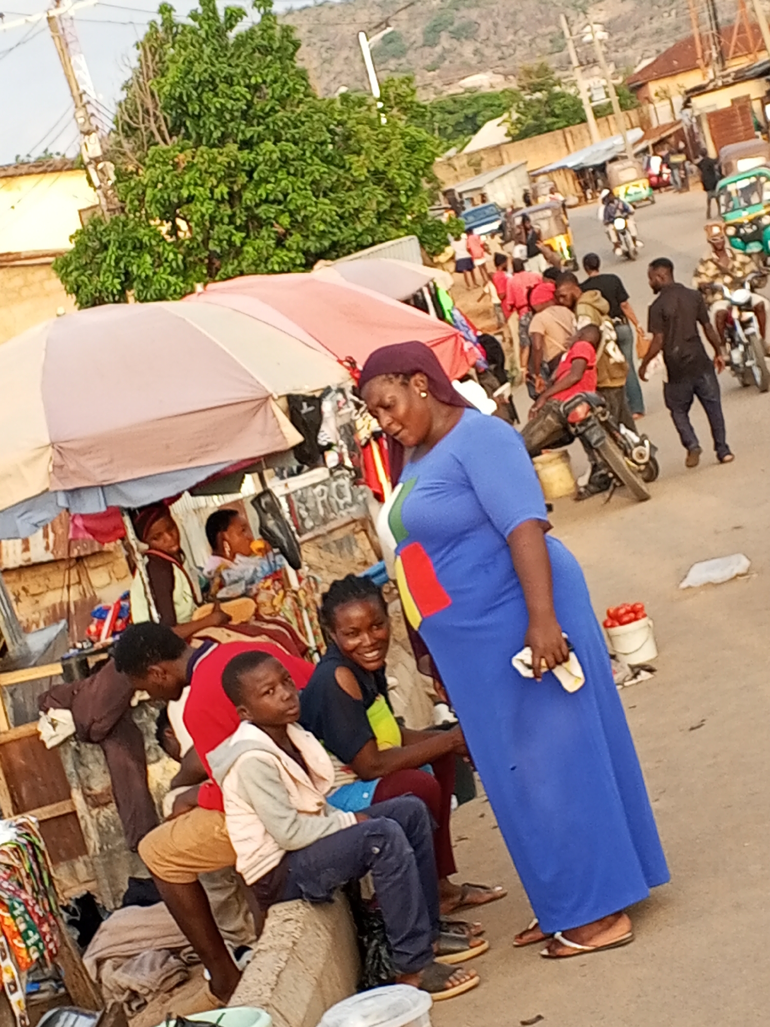 The image captures a street scene in Gwong, Nigeria. In the foreground, a woman wearing a blue dress and a headscarf stands on a paved road, holding what appears to be a white cloth or paper. Adjacent to her, several people are seated or leaning against what looks like a low concrete barrier or a makeshift seating area under an umbrella-covered stall. Two women and a young boy are facing the camera, while others are either partially visible or turned away. The stall appears to be selling various items.
In the background, the street is bustling with activity. There are people walking, motorcycles, and auto-rickshaws (tuk-tuks) navigating the street. Buildings and a hill are visible further in the background. The time of day appears to be late afternoon, given the angle of the sunlight. A bucket of tomatoes is to the woman's right.