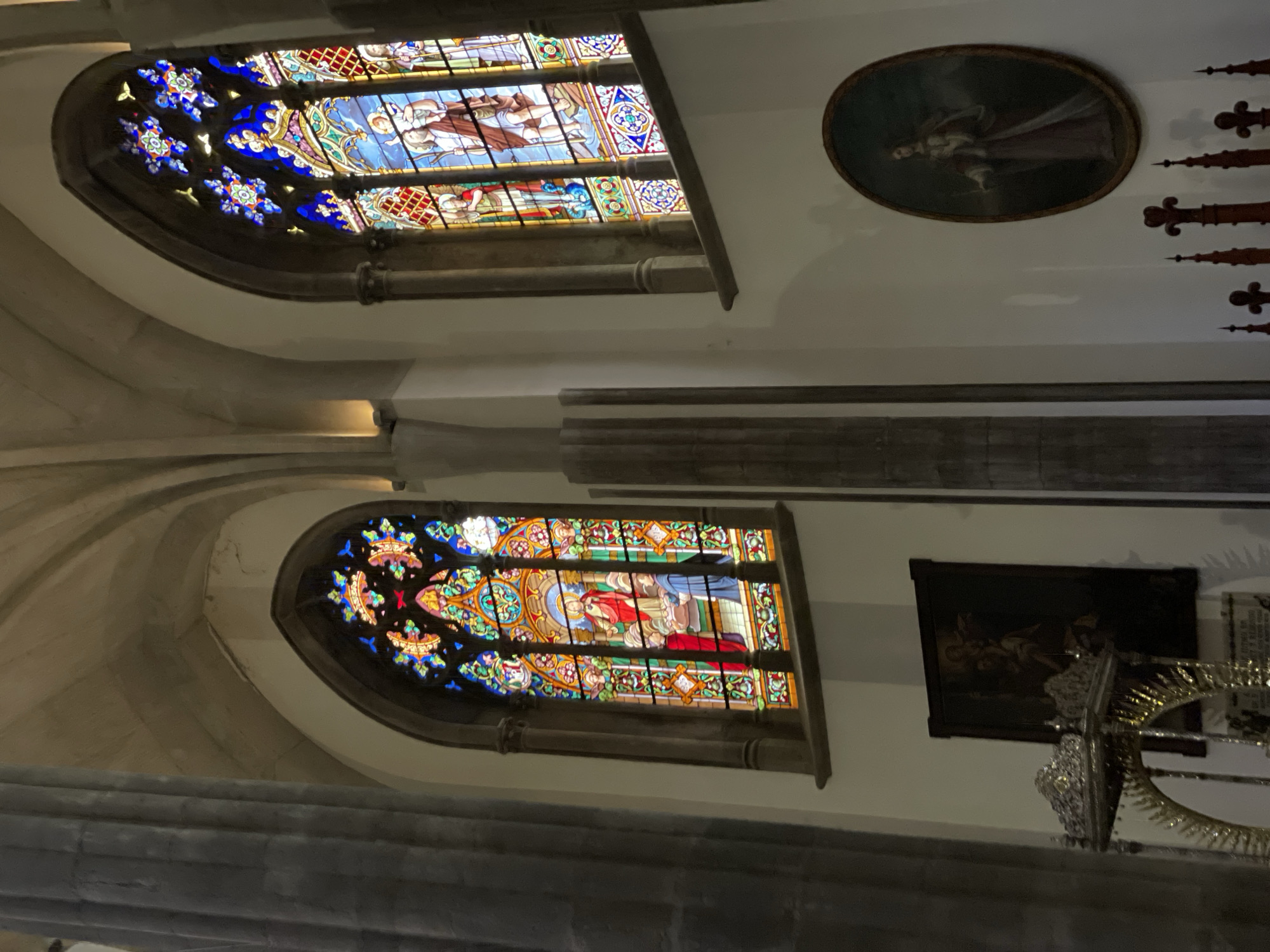 This media file captures the interior of a historic building, likely a church or cathedral, characterized by its architectural grandeur and religious art. The scene is dimly lit, with light primarily emanating from two large, ornate stained glass windows.
The upper section of the image features an arched stained glass window vibrant with blues, reds, greens, and yellows. It depicts religious figures, including a central figure with a halo, alongside other individuals. The upper parts of the arch are adorned with intricate floral or star-like patterns. To the right and slightly above this window, an oval-framed painting is visible on the light-colored wall. It portrays a solitary figure in flowing robes against a dark background, subtly highlighted by the ambient light. Further to the right, several dark, carved wooden spires or finials suggest decorative architectural elements.
A substantial dark beam or structural element spans horizontally across the middle of the image, dividing the upper and lower wall sections.
In the lower half, another large, arched stained glass window mirrors the style of the upper one, displaying vibrant religious narrative panels, with one figure prominently featured in a red cloak. To the right of this window, a rectangular painting is hung; it is very dark and indistinct, possibly depicting a multi-figured classical or religious scene. Below this painting, a highly ornate, gilded metal object with intricate scrollwork, possibly part of an altar or a large decorative fixture, reflects light.
The walls appear to be made of light-colored stone or plaster, providing a contrast to the darker architectural details and the glowing stained glass. The overall atmosphere conveys a sense of history, reverence, and artistic beauty. No specific activity or event is depicted, and no visible text is present. The setting is consistent with a religious building found in San Cristóbal De La Laguna, Spain.