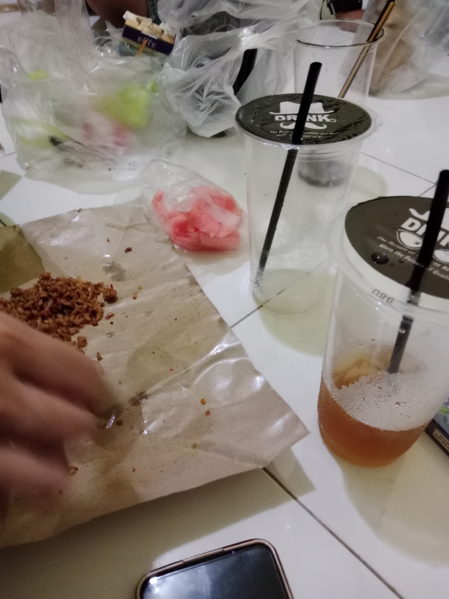 The image depicts a close-up, overhead view of a table at a casual eatery in West Jakarta, Indonesia. In the foreground, a hand is visible, reaching towards a small pile of fried shallots on a piece of brown paper. Next to the shallots is a clear plastic bag containing pink, sliced pickled radishes. To the right of these food items are two clear plastic cups with black lids and black straws, suggesting a beverage purchase. One cup is partially filled with a yellowish liquid and condensation. The background shows more plastic bags, some appearing to contain food items. The overall environment suggests a take-out or casual dining setting.