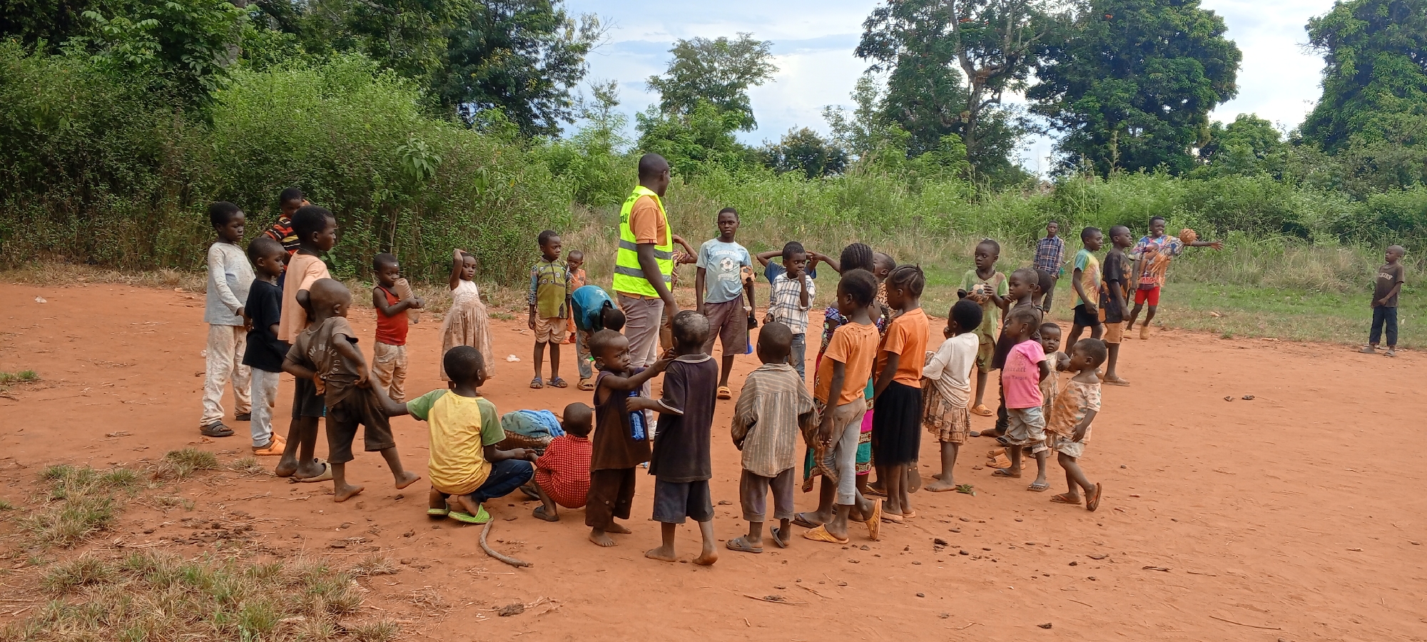 The scene depicts a group of approximately 30 children of various ages gathered in a dirt clearing in Ngalula, Democratic Republic of the Congo. The children are primarily dressed in simple clothing. Some are barefoot, while others wear sandals or shoes. An adult male wearing a bright orange shirt and a reflective yellow vest stands among the children. The children are grouped in the center of the image, while a couple of other children stand in the background. The backdrop consists of lush green vegetation, including trees and bushes. The ground is reddish-brown dirt, indicative of the soil found in the region.