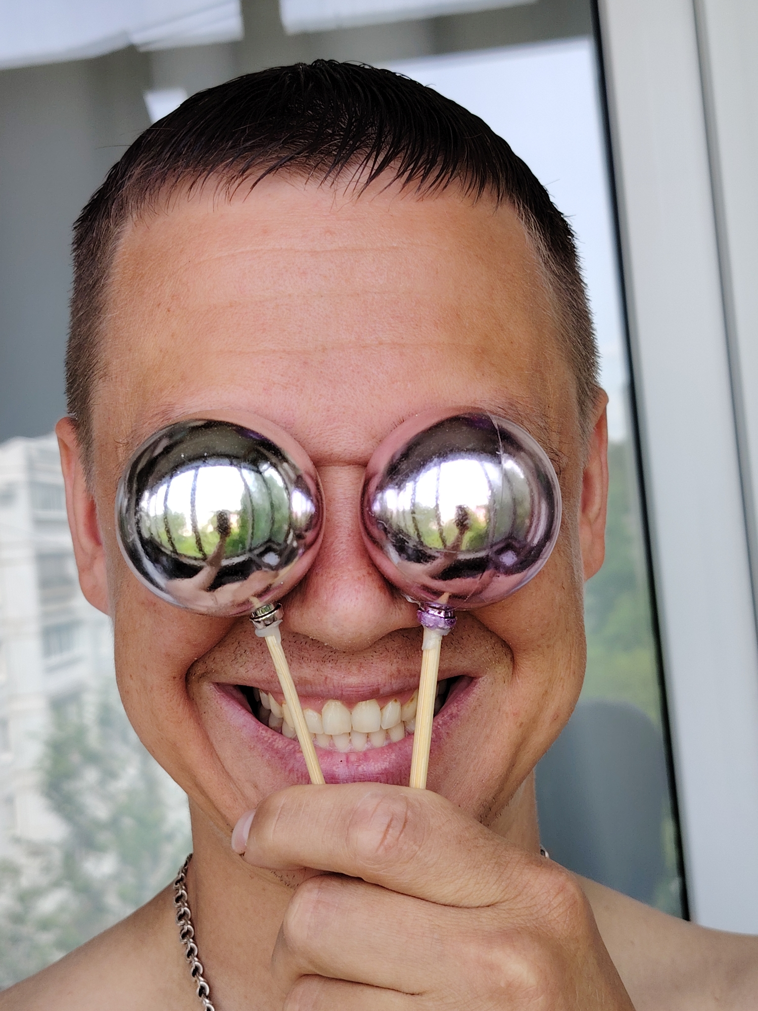 A close-up, waist-up shot features a man in front of a white wall, holding two reflective, spherical ornaments on sticks in front of his eyes, giving the impression of exaggerated eyeballs. He is grinning, showing his teeth, and the ornaments are positioned close to his nose. The man has short, dark hair and is wearing a silver chain necklace. Reflections on the ornaments show a building and greenery, indicating an outdoor setting. The scene is located in the city of Chaikovsky, Russia.