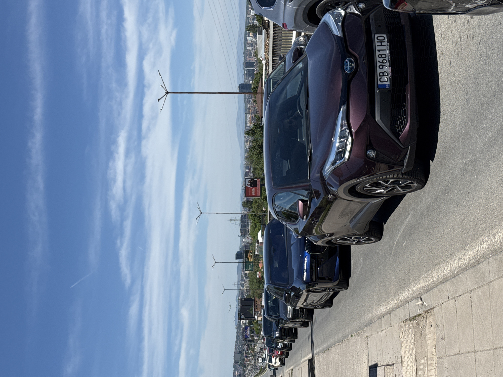 The image shows a row of parked cars on a street in Sofia, Bulgaria. The cars are parked along the curb on the right side of the frame. The foremost car is a dark maroon/purple Toyota C-HR with the license plate "CB 9681 HO." Other cars in the row are various dark colors, including blue and black. In the background, a cityscape of Sofia is visible, with buildings, trees, and mountains. Utility poles line the street with their lights. The sky is blue with some clouds. The perspective is taken from a standing point on the sidewalk, potentially on the high street.