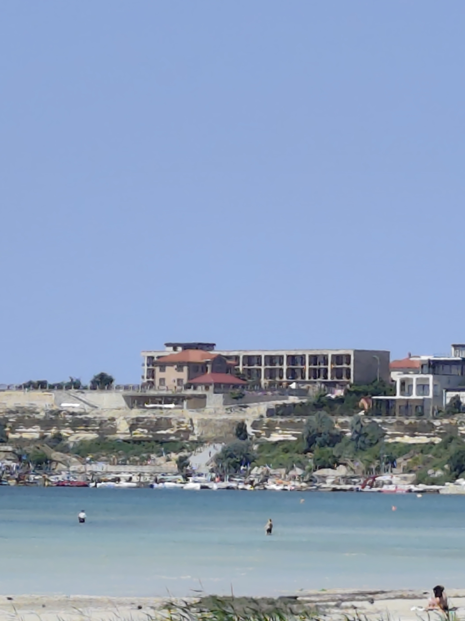 The image depicts a coastal scene with buildings situated on a cliff overlooking a body of water. The buildings appear to be a hotel or resort complex, with multiple stories and balconies. The cliff face is rugged and shows signs of erosion, with vegetation growing in patches. Below the cliff, a shoreline is visible, lined with numerous small boats and what appear to be kiosks or stalls. The water is a clear blue, with some ripples indicating a light breeze. A few individuals are visible in the water, wading or swimming. In the foreground, some dry vegetation is present on a sandy area. The sky is a clear, bright blue, suggesting a sunny day. This scene is consistent with the coastal areas of Aktau, Kazakhstan, known for its beaches and resort developments along the Caspian Sea.