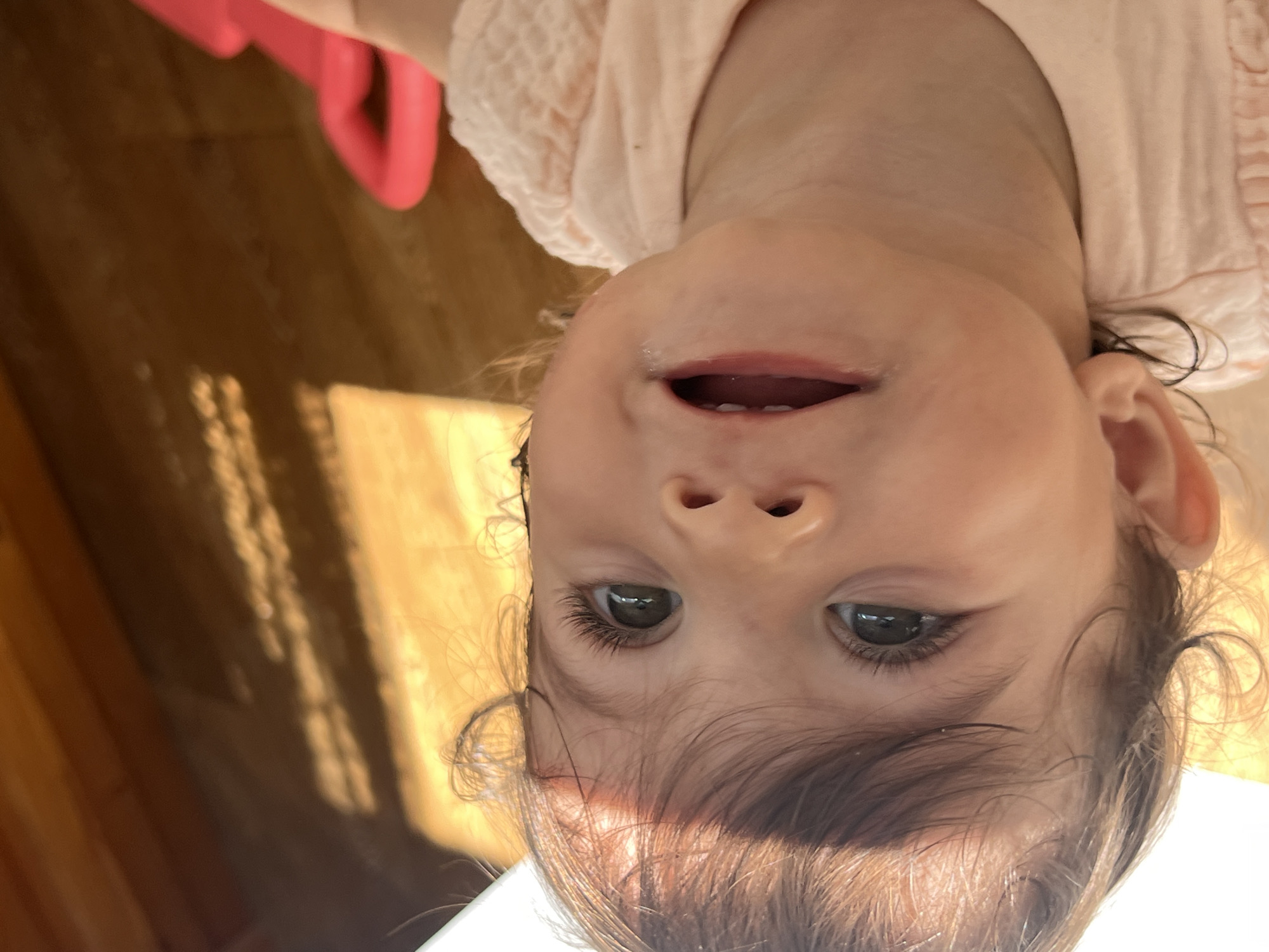 The image is a close-up, low-angle, portrait-style photograph of a young child. The child, with dark hair and green eyes, is wearing a light pink, ruffled garment. The child's mouth is open, and they appear to be looking upwards towards the camera. Sunlight is casting shadows across the wooden floor in the background, where a bright pink object is partially visible. The scene is indoors, likely a home environment. The location is not specified within the image.