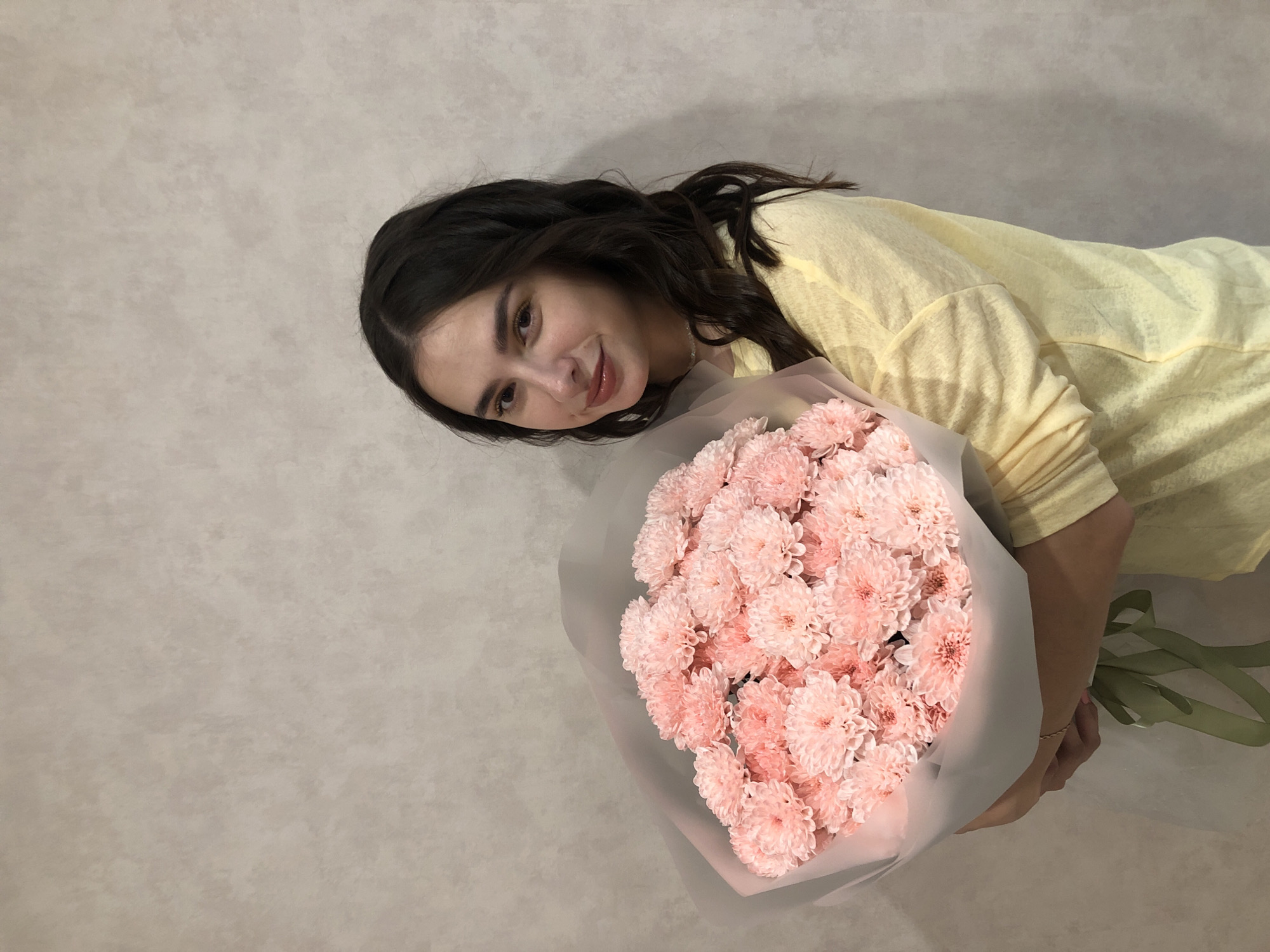 A young woman with dark, wavy hair and brown eyes is standing against a neutral, textured wall. She is wearing a yellow long-sleeved shirt and is holding a large bouquet of light pink chrysanthemums wrapped in light grey paper and tied with a green ribbon. Her hands are visible at the bottom of the bouquet, supporting it. The woman is looking towards the camera with a slight smile. The background is a plain, light-colored wall. The lighting is soft, illuminating the subject and the flowers. This image was taken indoors.