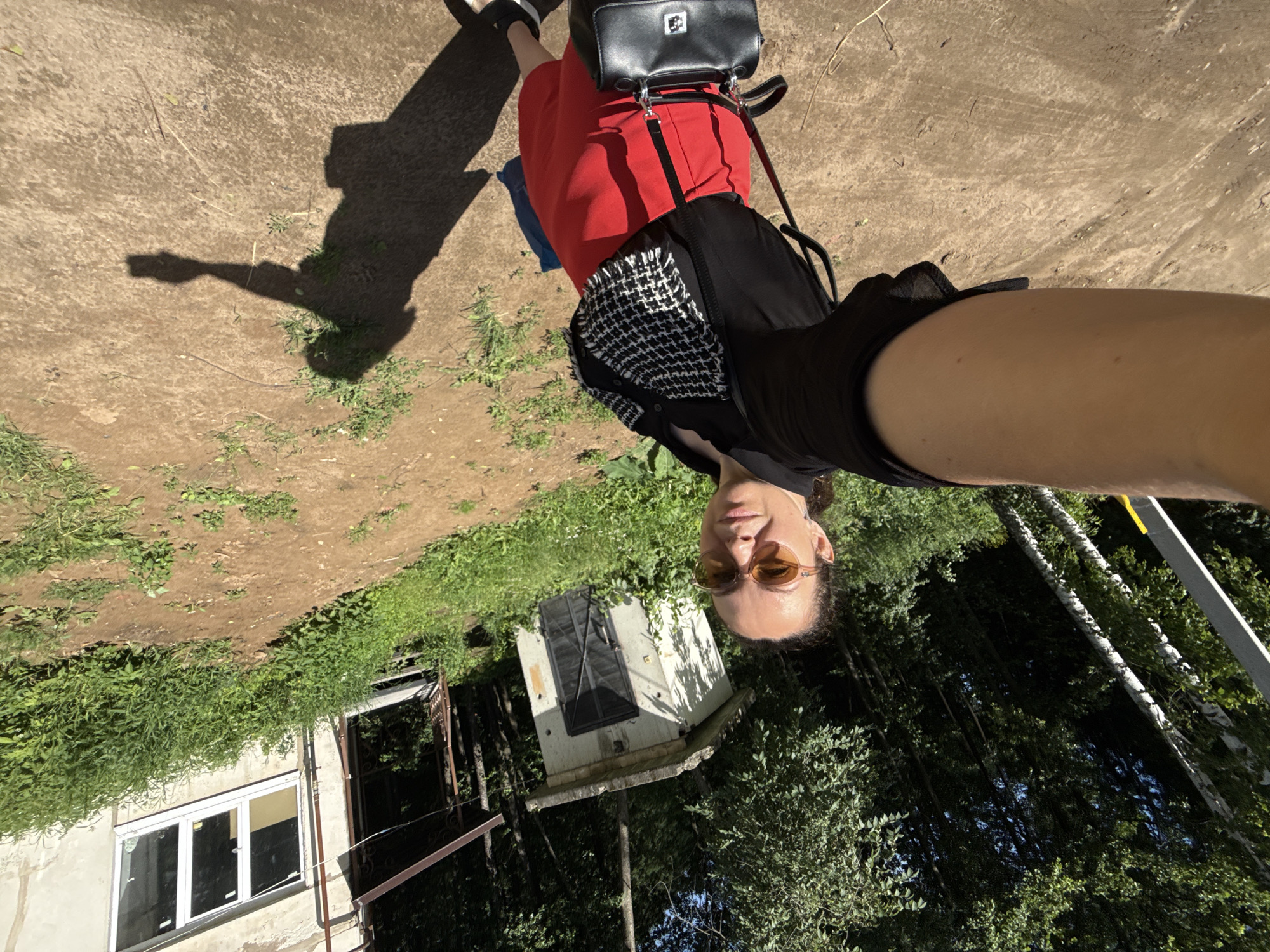 In Chaikovsky, Russia, a person with sunglasses and a black shirt is taking a selfie outdoors. The person is wearing a red skirt and a patterned scarf over their shoulders. A black purse is visible next to them. The background shows a building, trees, and a dirt path. The sun casts a shadow of the person and their arm, holding a phone, onto the path.