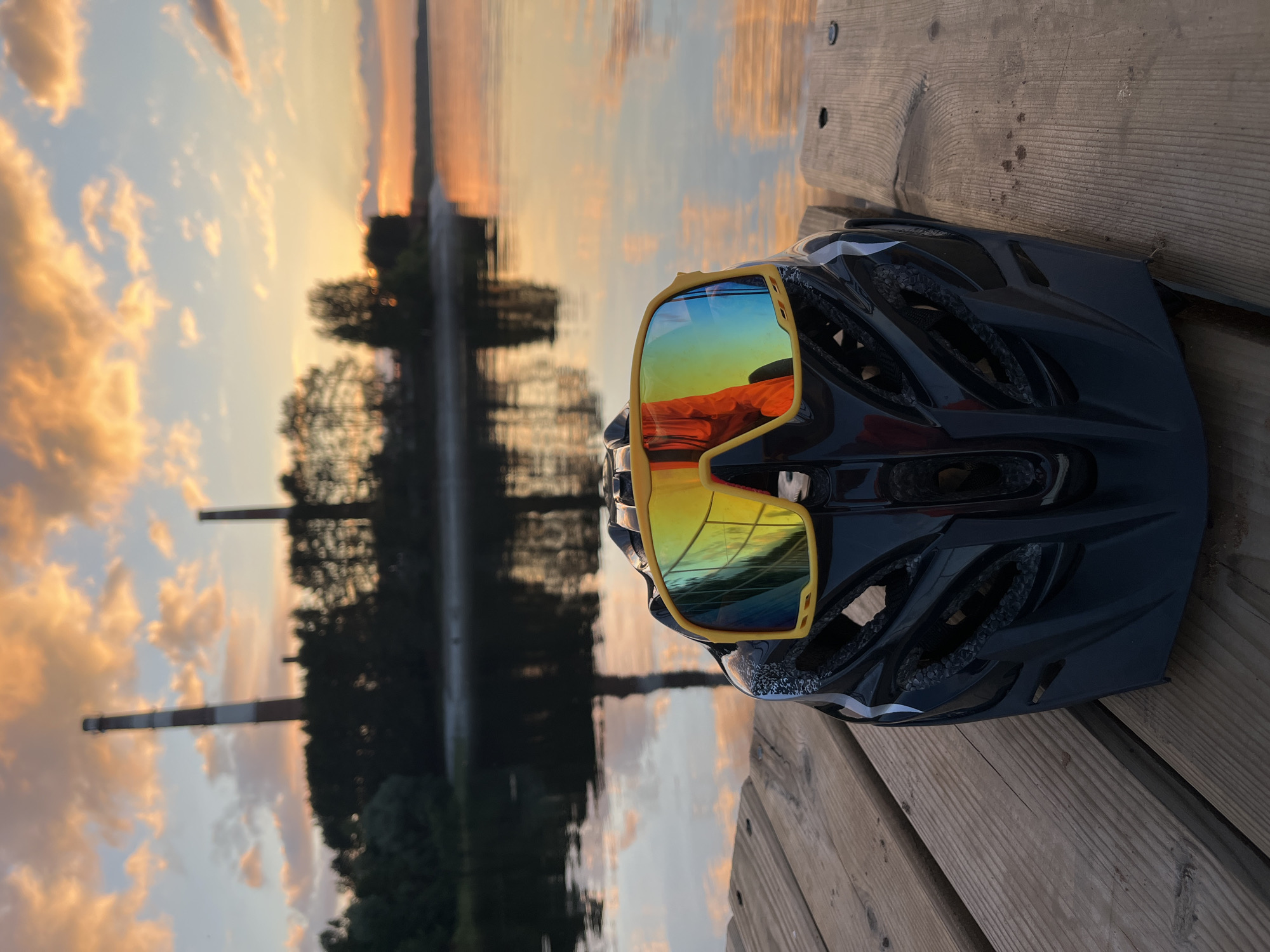 A black bicycle helmet with yellow-rimmed cycling glasses rests on a wooden dock. The glasses have mirrored lenses reflecting a sunset with orange and yellow clouds, and a body of water. In the background, a calm body of water is visible, reflecting the sky and silhouetted trees. Two tall smokestacks, one with red and white bands, stand on the far side of the water, suggesting an industrial presence. The scene is bathed in the warm light of a sunset, indicating it is late evening. The overall mood is peaceful and serene, possibly after a day of cycling or outdoor activity. There is no visible text in the image.