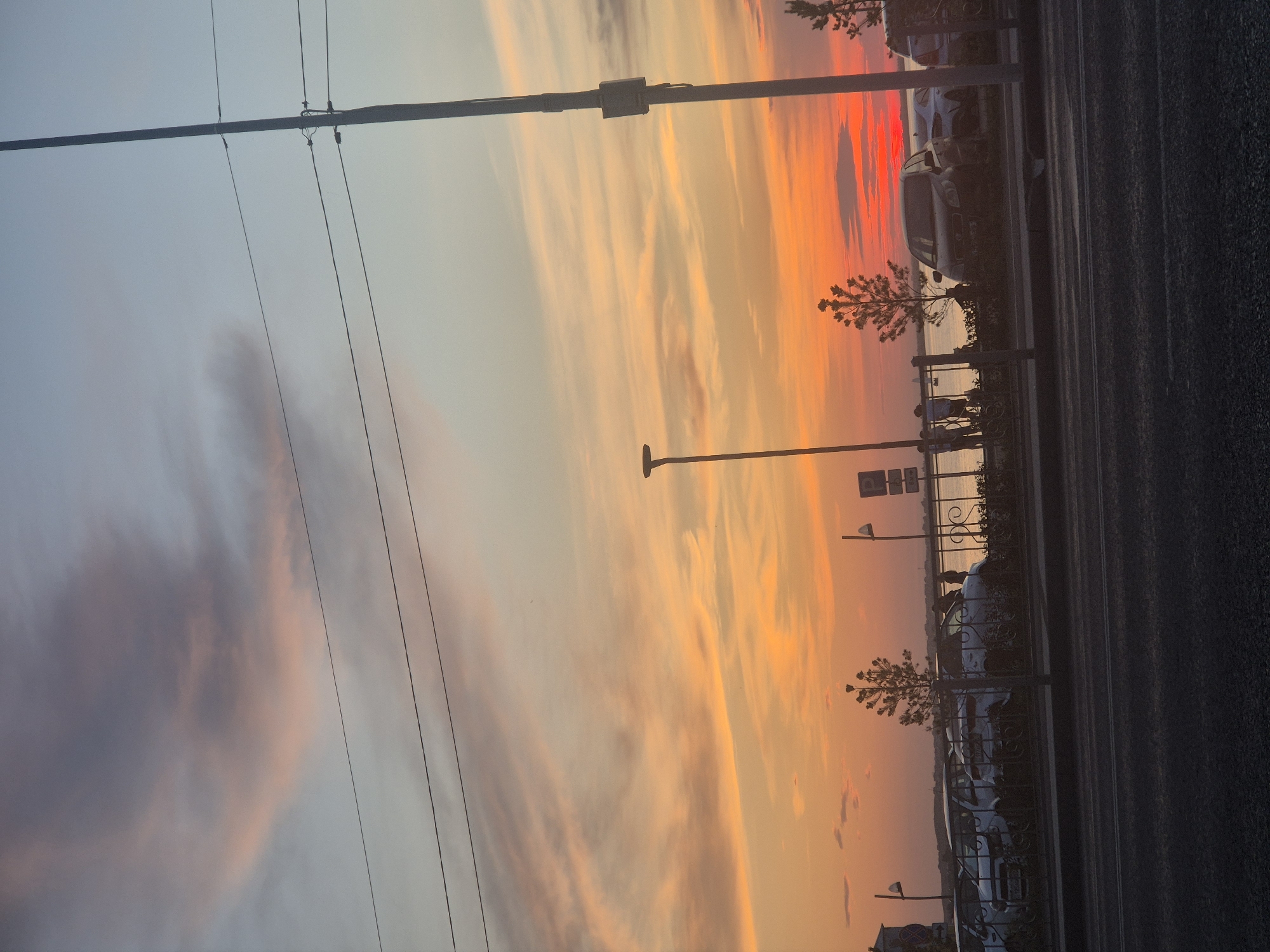 The image captures a sunset over a body of water, likely Lake Borovoe, in Kokshetau, Kazakhstan. The sky is a vibrant spectacle of orange and pink hues, with streaks of clouds painted across the canvas. In the foreground, a road is visible, with parked cars lining the other side of the street. A fence separates the road from a promenade where several individuals are seen standing and walking, silhouetted against the colorful sky. Streetlights and utility poles punctuate the scene, adding a touch of urban infrastructure to the natural beauty of the sunset. The overall atmosphere is serene, with the warm glow of the setting sun casting a peaceful ambiance over the landscape.