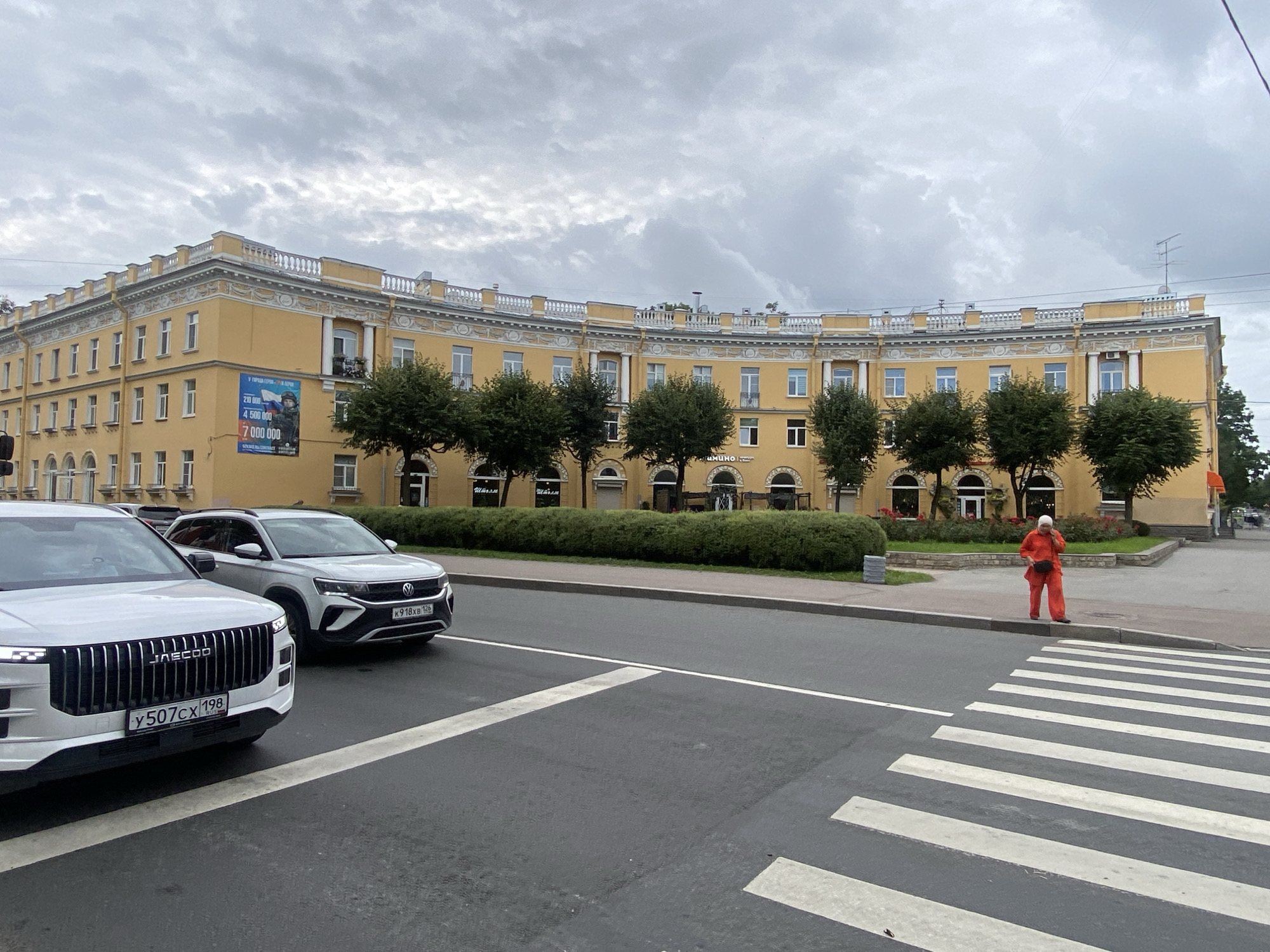 An overcast day in Pushkin, Russia, with a yellow, curved building in the background. The building has ornate white trim and several windows. A large banner featuring a soldier is visible on the left side of the building. In the foreground, two white cars are parked on the street. A person wearing a bright orange outfit is standing on the sidewalk near a pedestrian crossing. The street is paved with asphalt, and there are white lines marking the pedestrian crossing. Trees and bushes line the sidewalk in front of the building.