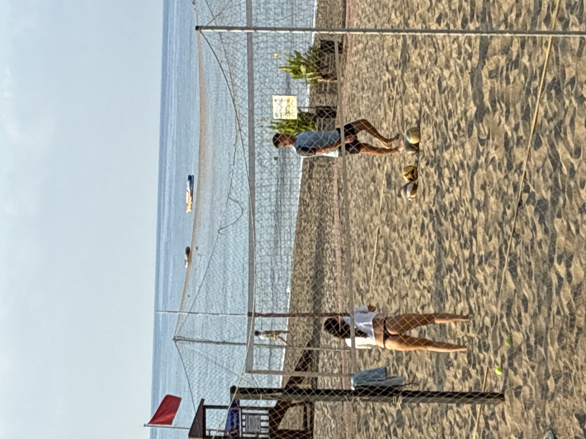 This image depicts a beach volleyball scene on a sunny day in Alanya, Turkey, with the Mediterranean Sea in the background. A red flag is visible on a pole to the left, indicating sea conditions. A volleyball net divides the sandy court where two individuals are present. In the foreground, on the left side of the net, a person wearing a white top and dark bikini bottoms stands facing away from the camera, appearing to be playing volleyball. On the right side of the net, a person in a light blue t-shirt and dark shorts is also on the court, looking towards the other player. Several volleyballs are scattered on the sand, and two tennis balls are near the person on the left. In the background, the sea is calm, with a few boats visible on the water. A lifeguard station or wooden structure is partially visible on the far left. The overall atmosphere is one of recreation and leisure on a coastal resort.