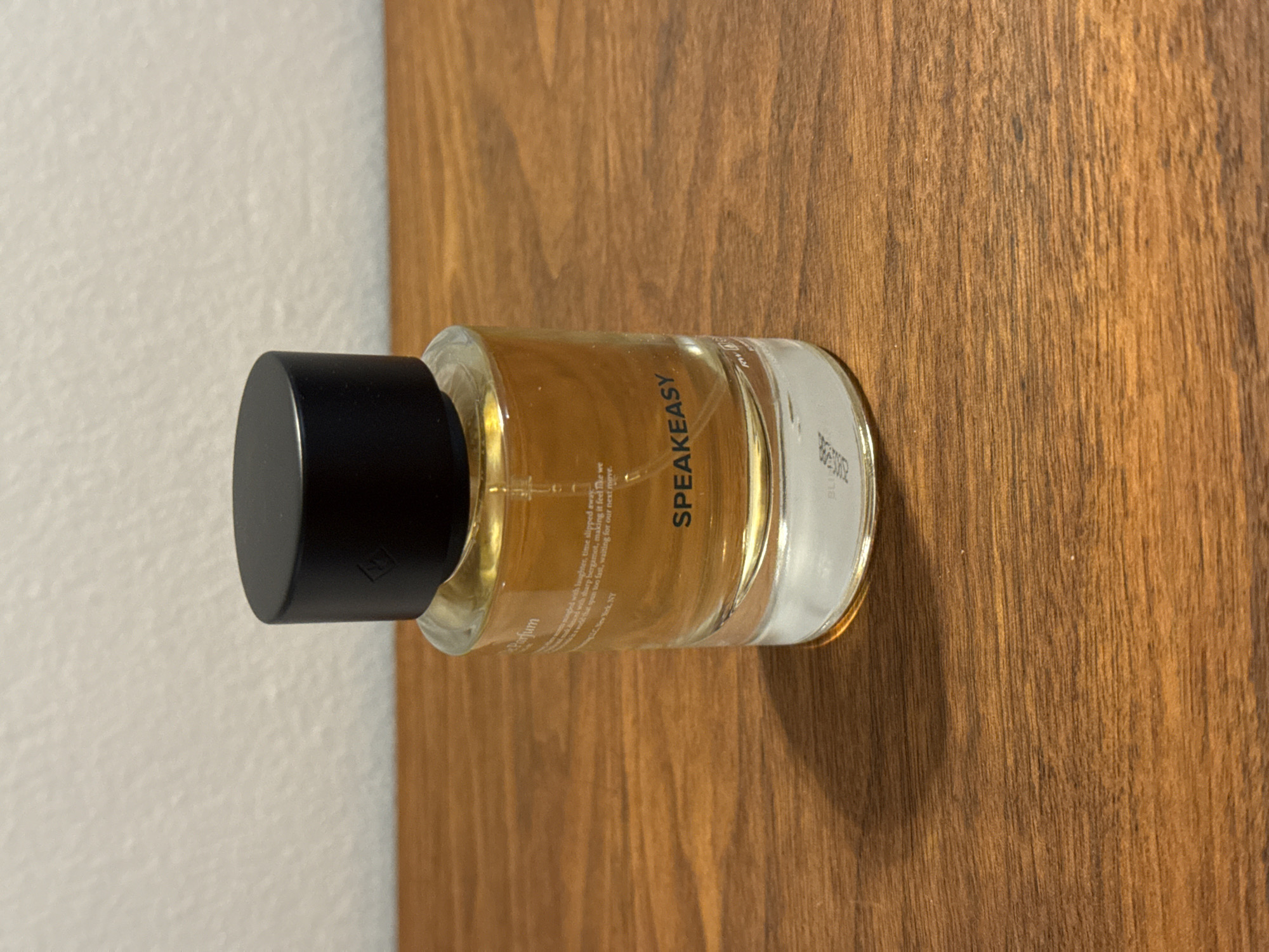 The image shows a bottle of "Speakeasy" parfum on a wooden surface. The bottle has a black cap and a clear glass body containing a golden liquid. Text on the bottle includes "Parfum" and a description referencing laughter and bergamot. The wooden surface exhibits a distinct grain pattern. The location in the city of Montreal, Canada cannot be determined from the image.