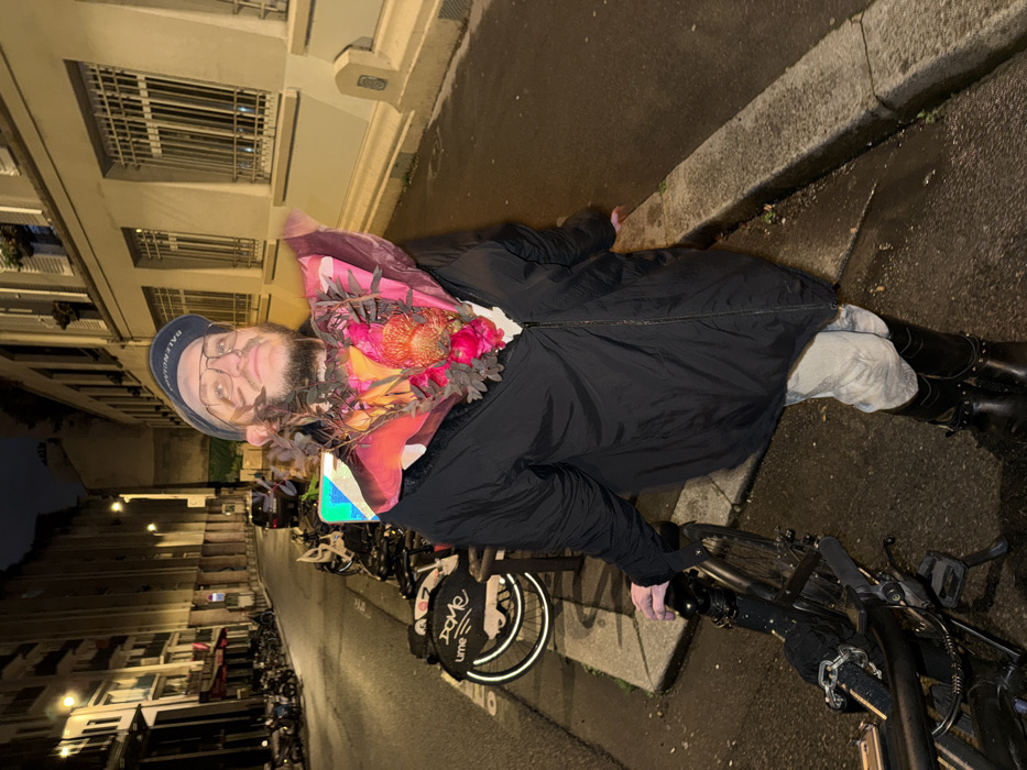 A person with a beard, glasses, and a "Balenciaga" cap stands on a street next to a bicycle. The person wears a black coat, light grey pants, and black boots with metal accents. They hold a bouquet of flowers wrapped in a colorful scarf with a floral pattern. Rows of parked bicycles are visible along the street, with European-style buildings in the background under a night sky. The image appears to have been rotated counter-clockwise.