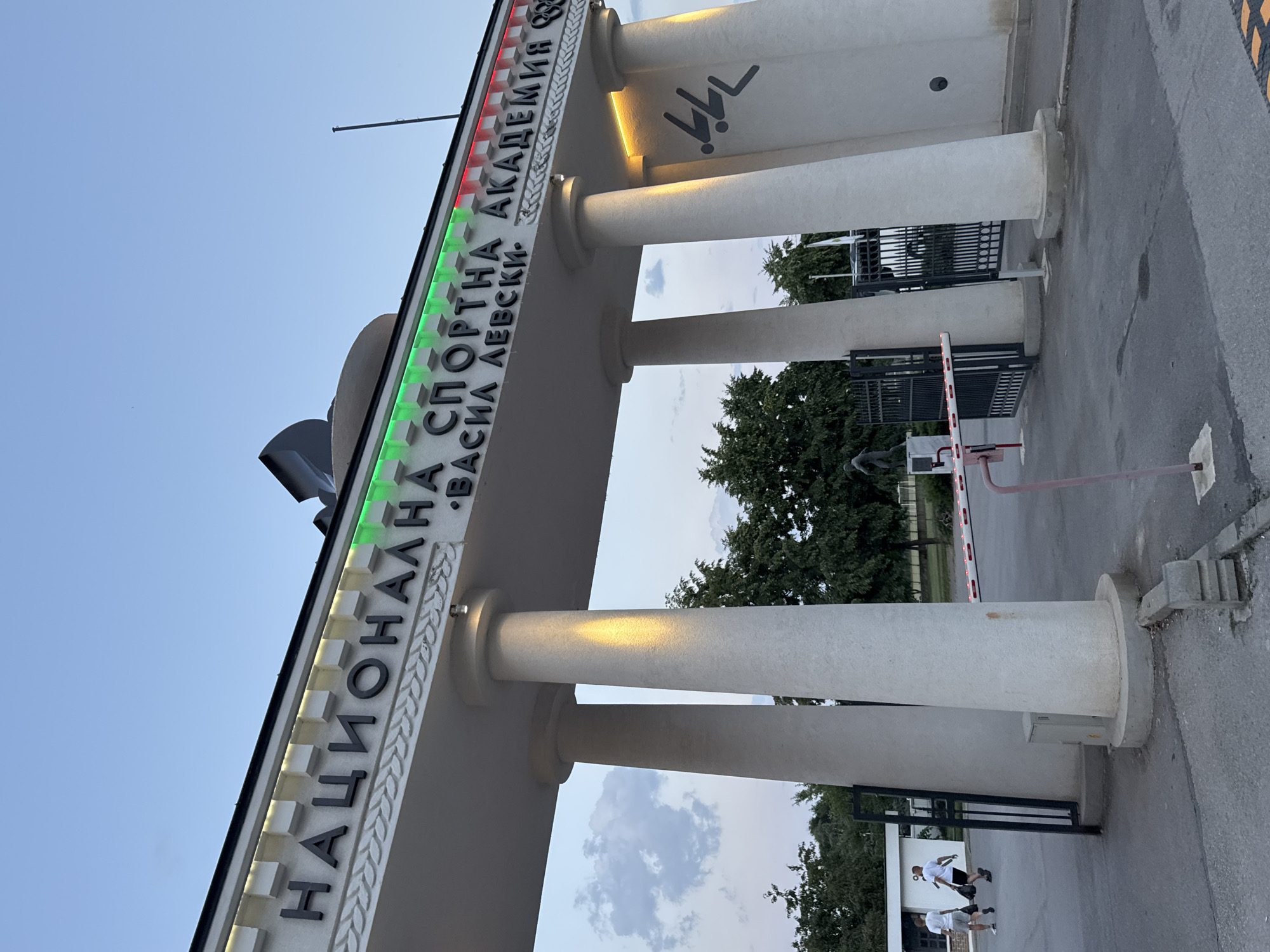 The image shows the entrance gate of the National Sports Academy "Vasil Levski" in Sofia, Bulgaria. The gate features a white archway with Cyrillic lettering that reads "НАЦИОНАЛНА СПОРТНА АКАДЕМИЯ ВАСИЛ ЛЕВСКИ". The lettering is accented by green and red lights, resembling the Bulgarian flag. Two large white columns support the archway. To the left of the gate, two individuals wearing white t-shirts and shorts are walking away from the camera, carrying black bags. Behind them, a white building and a black metal fence are visible. In the middle of the frame, a red and white striped barrier is lowered across the entrance. The sky is overcast, and lush green trees are visible in the background. The overall scene depicts a structured environment, likely during the evening, with a focus on the monumental entrance of the academy.