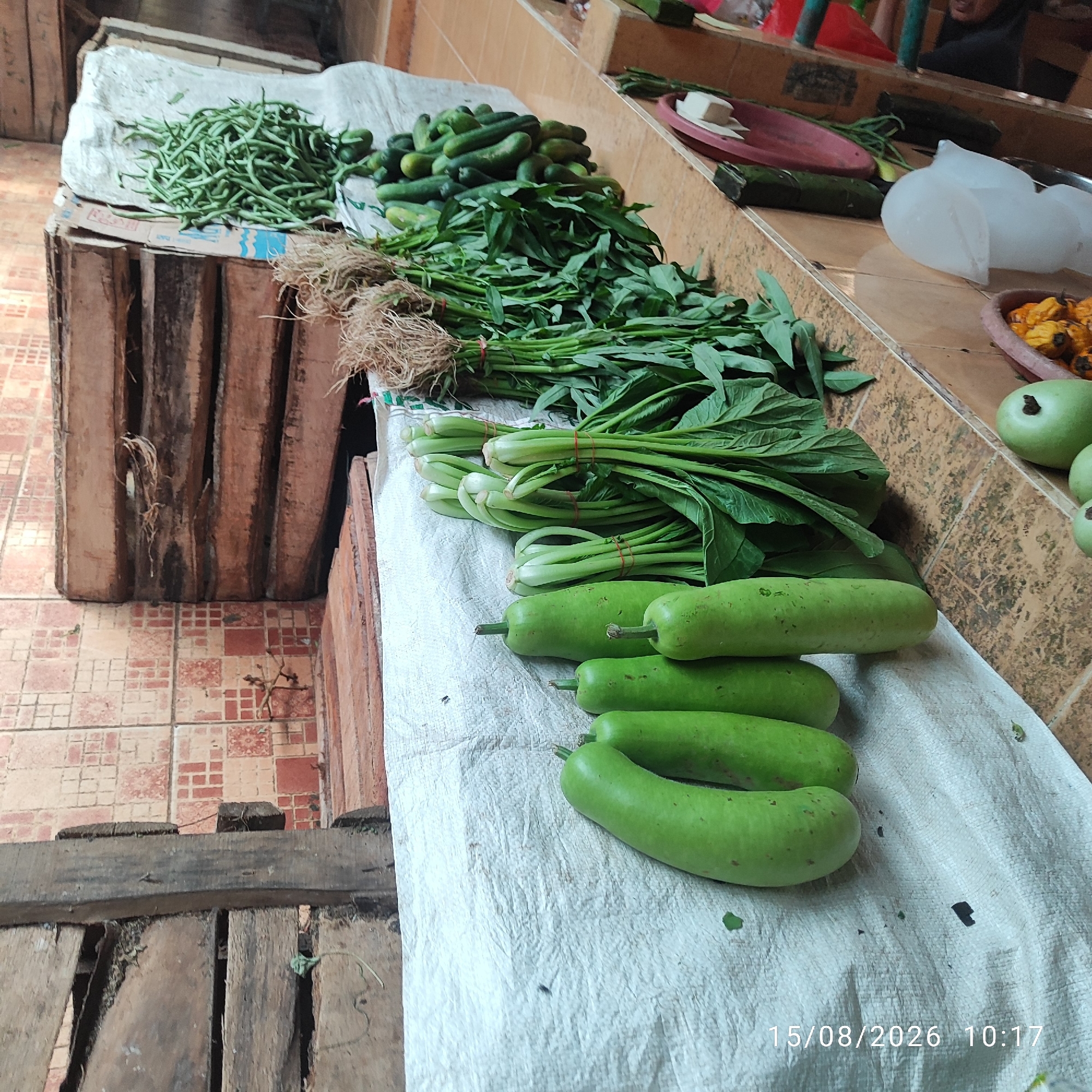 A street market stall in Pembaharuan, Indonesia, displays a variety of fresh produce. The stall is covered with a white cloth. In the foreground, several green gourds are neatly arranged. Behind them, bunches of leafy green vegetables, possibly water spinach, are tied with red elastic bands, their roots exposed. Further back, a pile of green beans and cucumbers are visible. The stall itself is made of weathered wood, with a tiled floor in the background. A digital timestamp in the bottom right corner reads "15/08/2026 10:17".