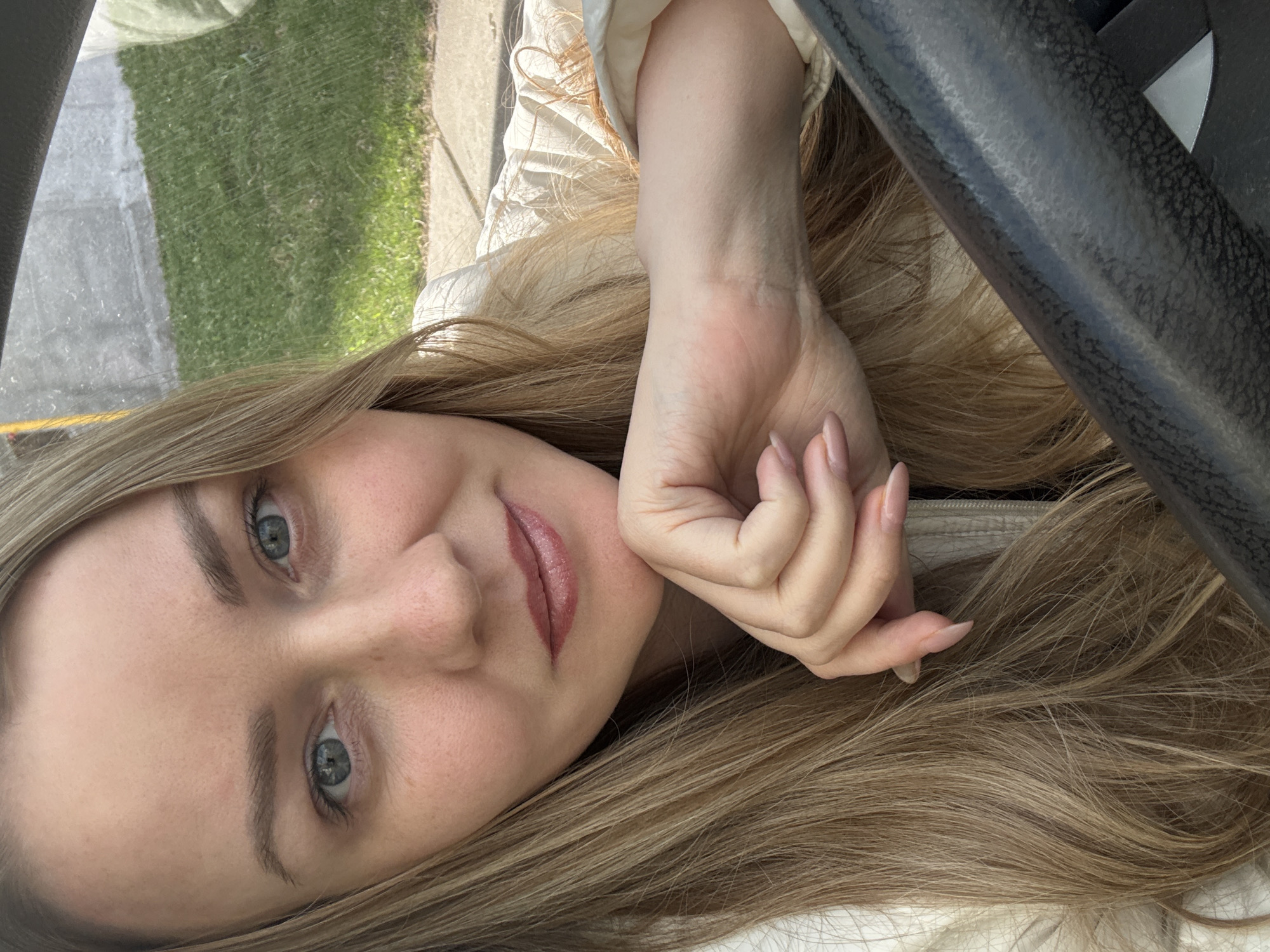 A young woman with blonde hair and light blue eyes is in a vehicle in Chaikovsky, Russia. Her head is tilted to the side, and her hand is near her face. A black steering wheel is visible to the right. Outside the window on the left is grass and a paved area. The woman is wearing a white top.