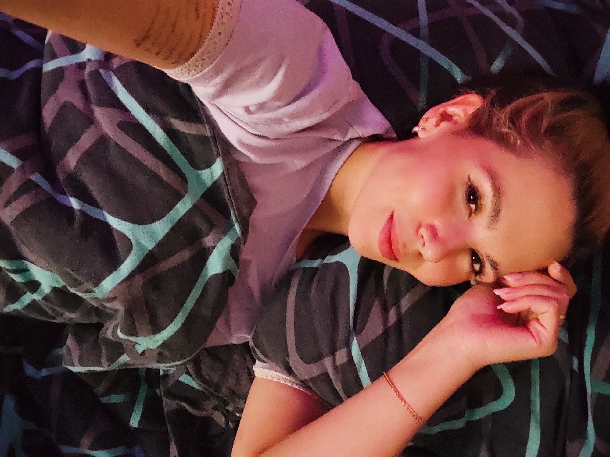 A woman is depicted in a close-up, high-angle shot, lying down indoors, likely on a bed, in Chaikovsky, Russia. She is partially covered by a dark blanket or duvet featuring a bold geometric pattern of teal and purple lines. The woman is wearing a light-colored, possibly lavender or pale purple, short-sleeved top with delicate lace trim visible on the sleeve and neckline.
Her head is resting, with her right arm bent and her hand gently touching her face near her chin. She has brown hair, pulled back or up, and is wearing visible eye makeup and red lipstick. A small, silver-colored hoop earring is noticeable in her right ear, and a thin, golden-colored bracelet adorns her left wrist. The lighting has a soft, warm quality, casting a slight pinkish-purple hue on parts of her face and shirt, suggesting either artificial lighting or a specific light source. She looks calmly towards the camera, conveying a relaxed demeanor. There is no visible text in the image.