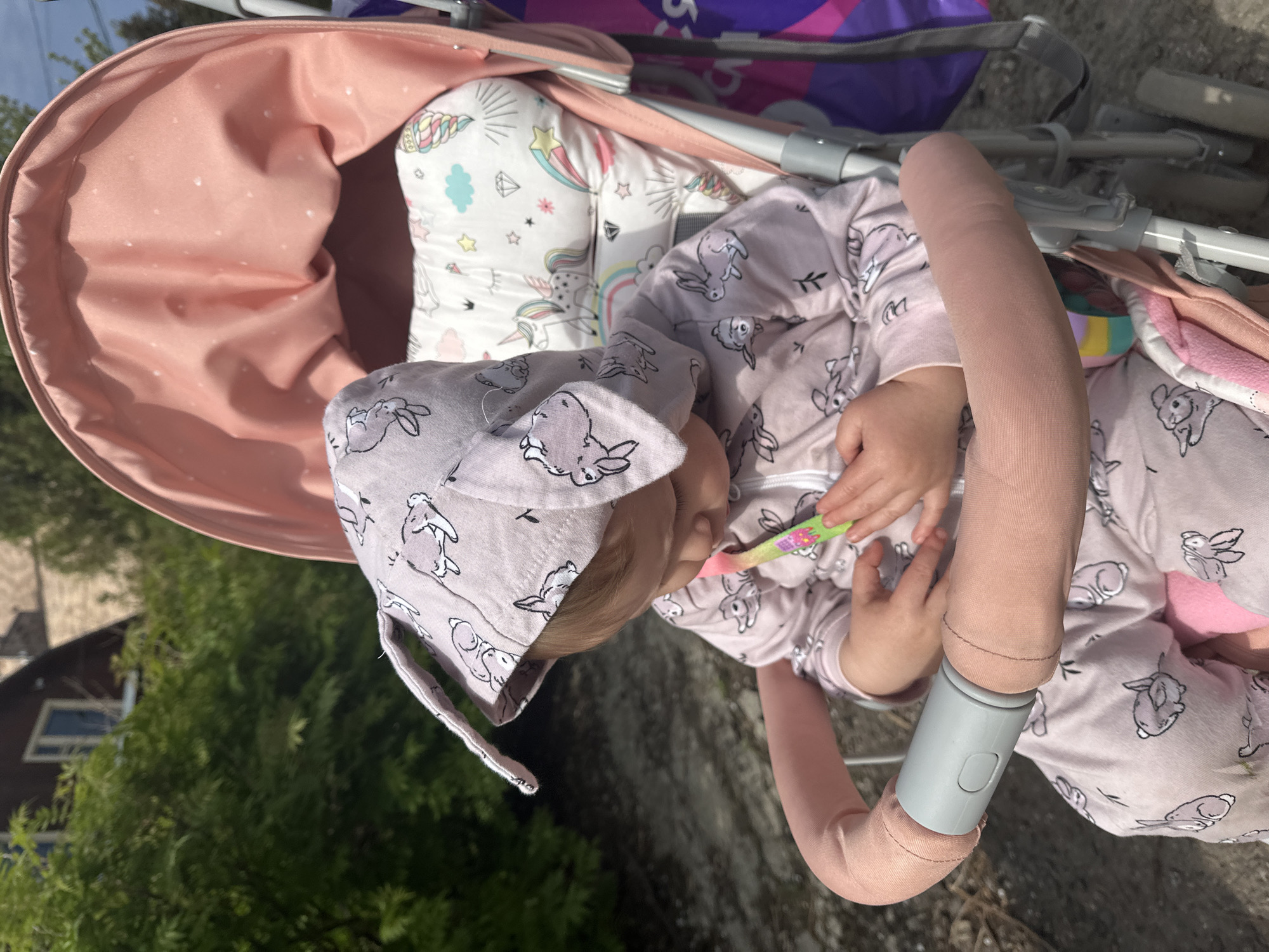 A young child wearing a pink bunny-patterned outfit and hat is lying in a pink stroller. The child is holding a pink and green object. The stroller has a white pillow with unicorn and rainbow designs. The background includes green foliage and a building with dark roofing, suggesting an outdoor urban or suburban environment. The specific location in Nizhny Novgorod, Russia, cannot be determined from this image.